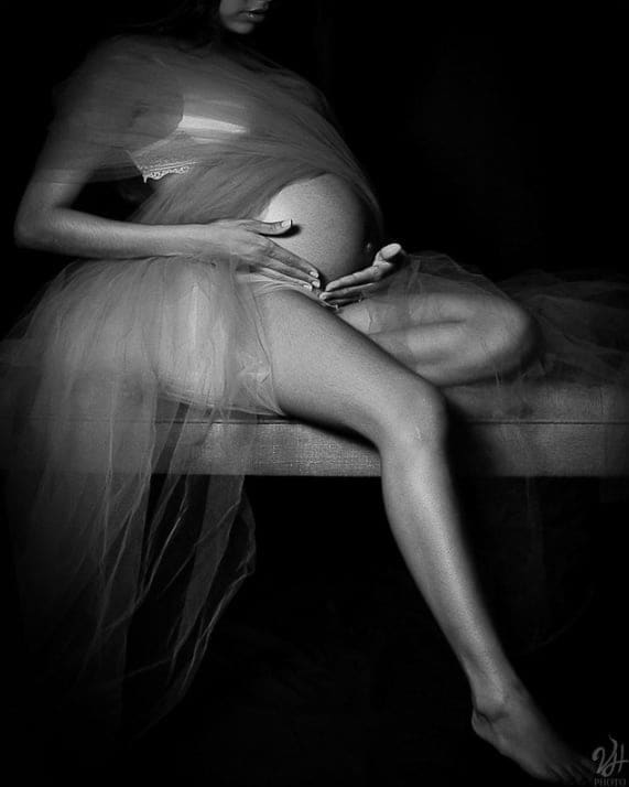 Scatto artistico in bianco e nero di una donna incinta con tulle trasparente seduta su una panca, che accarezza il pancione.