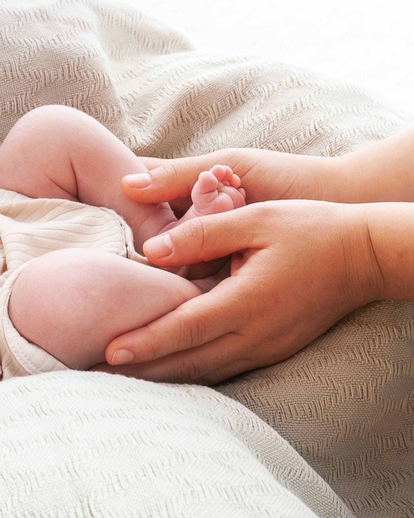 Piccolo piede di un neonato stretto tra le mani di un adulto, su coperte color panna in luce chiara e morbida.