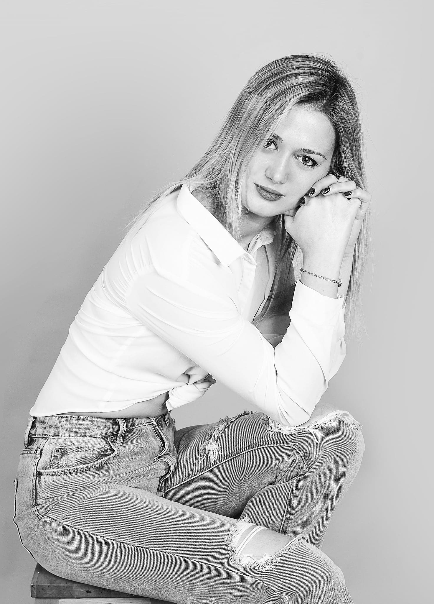 In bianco e nero, ragazza bionda in camicia bianca e jeans strappati seduta con la testa appoggiata alle mani, su fondo chiaro