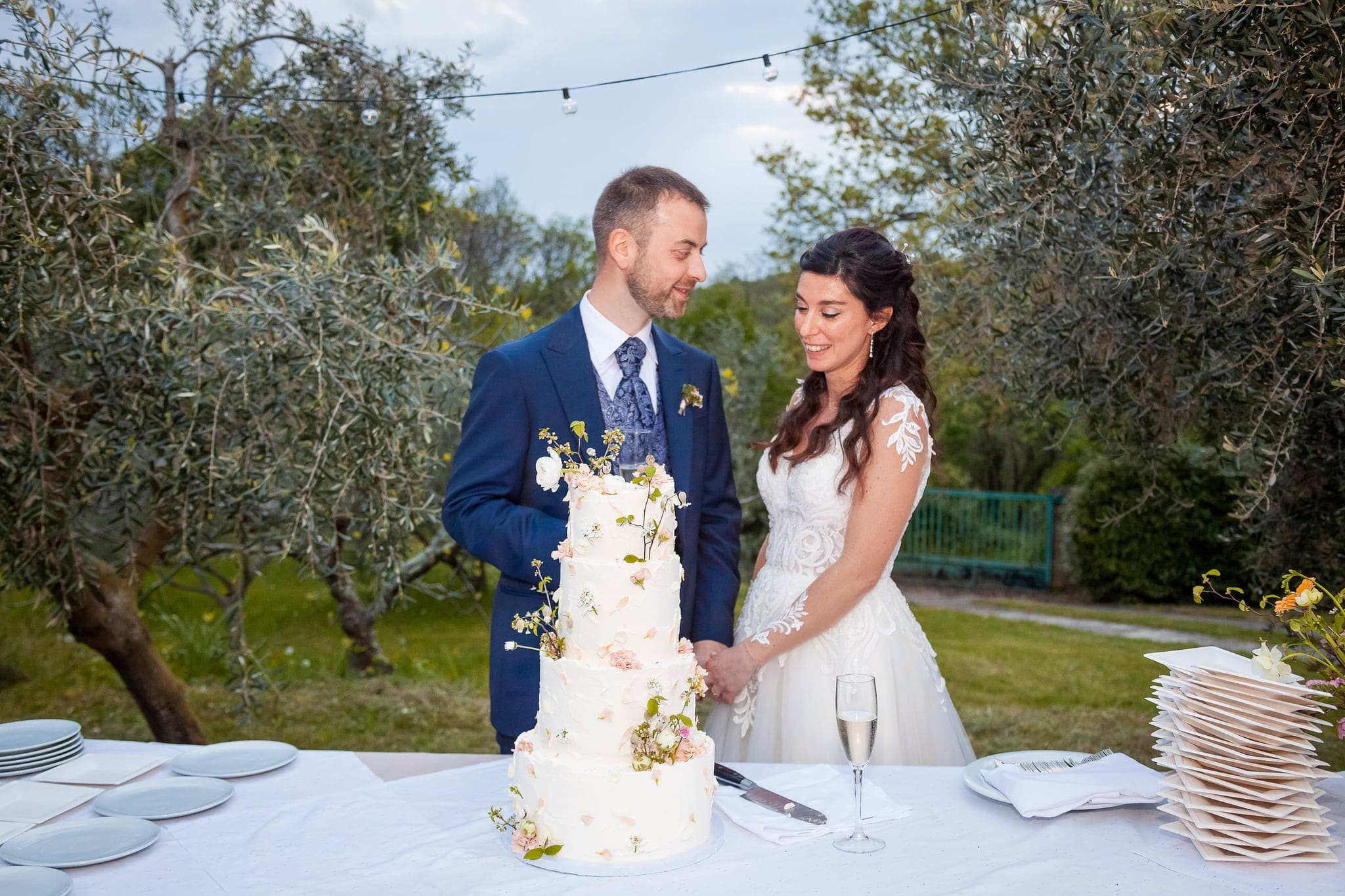 Sposi che tagliano la torta nuziale a più piani decorata con fiori, tra gli ulivi, matrimonio sulla Terra del Carso