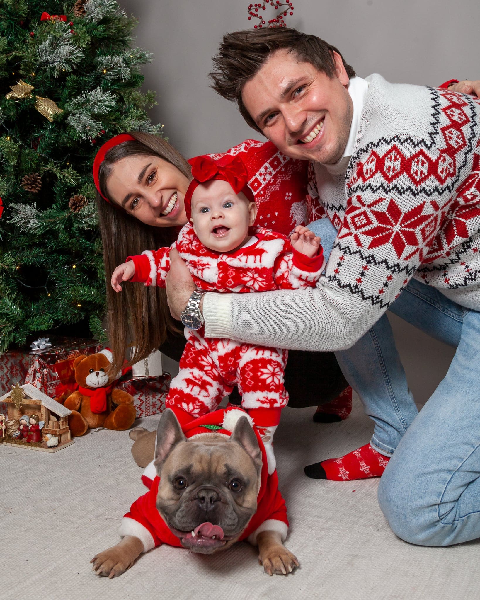 Papà, mamma e neonata in tuta natalizia sorridono vicini, mentre il bulldog francese in costume rosso spunta in primo piano