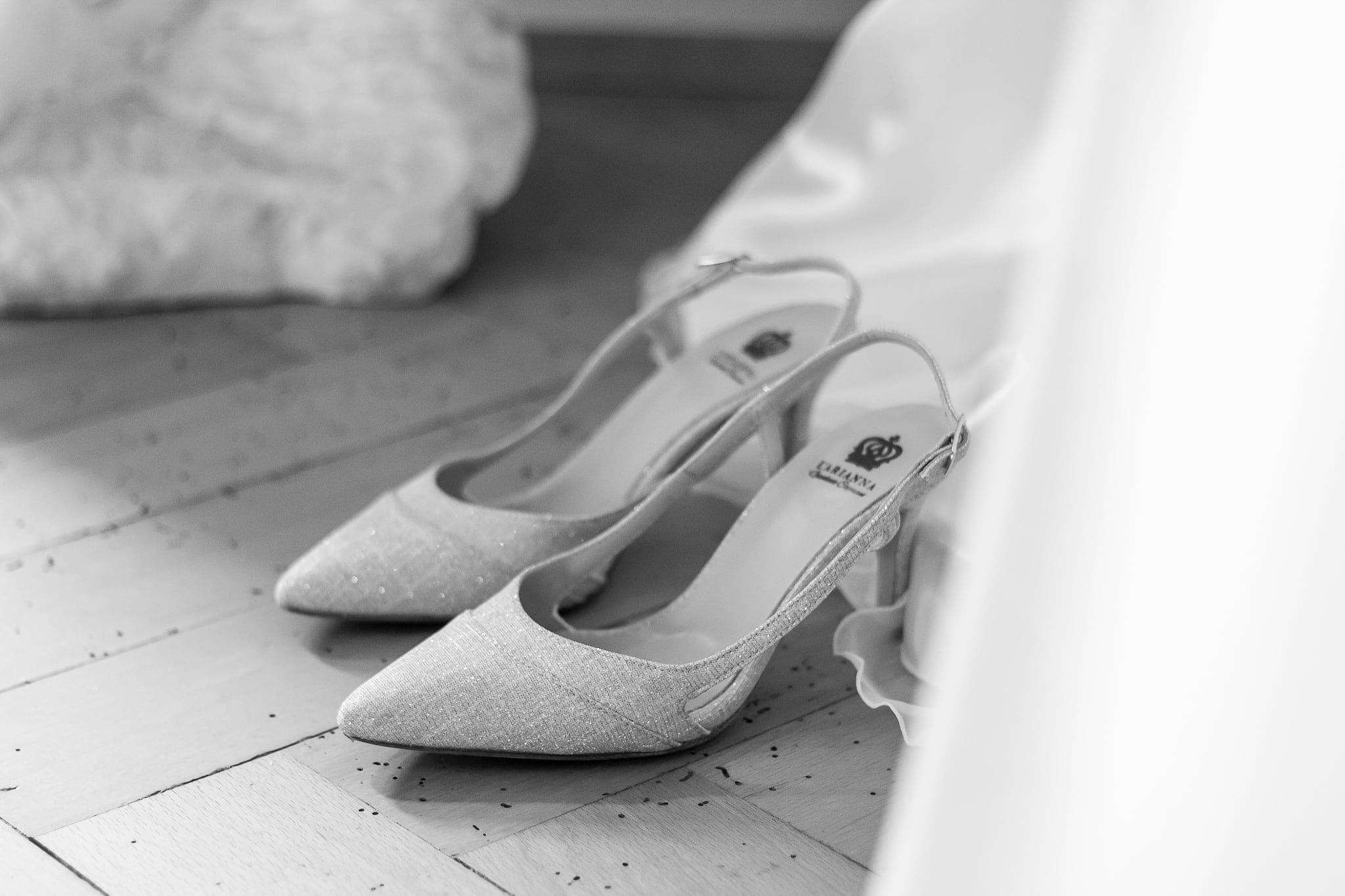 Scarpe da sposa con tacco e cinturino appoggiate sul pavimento accanto all'abito, in bianco e nero, durante la vestizione