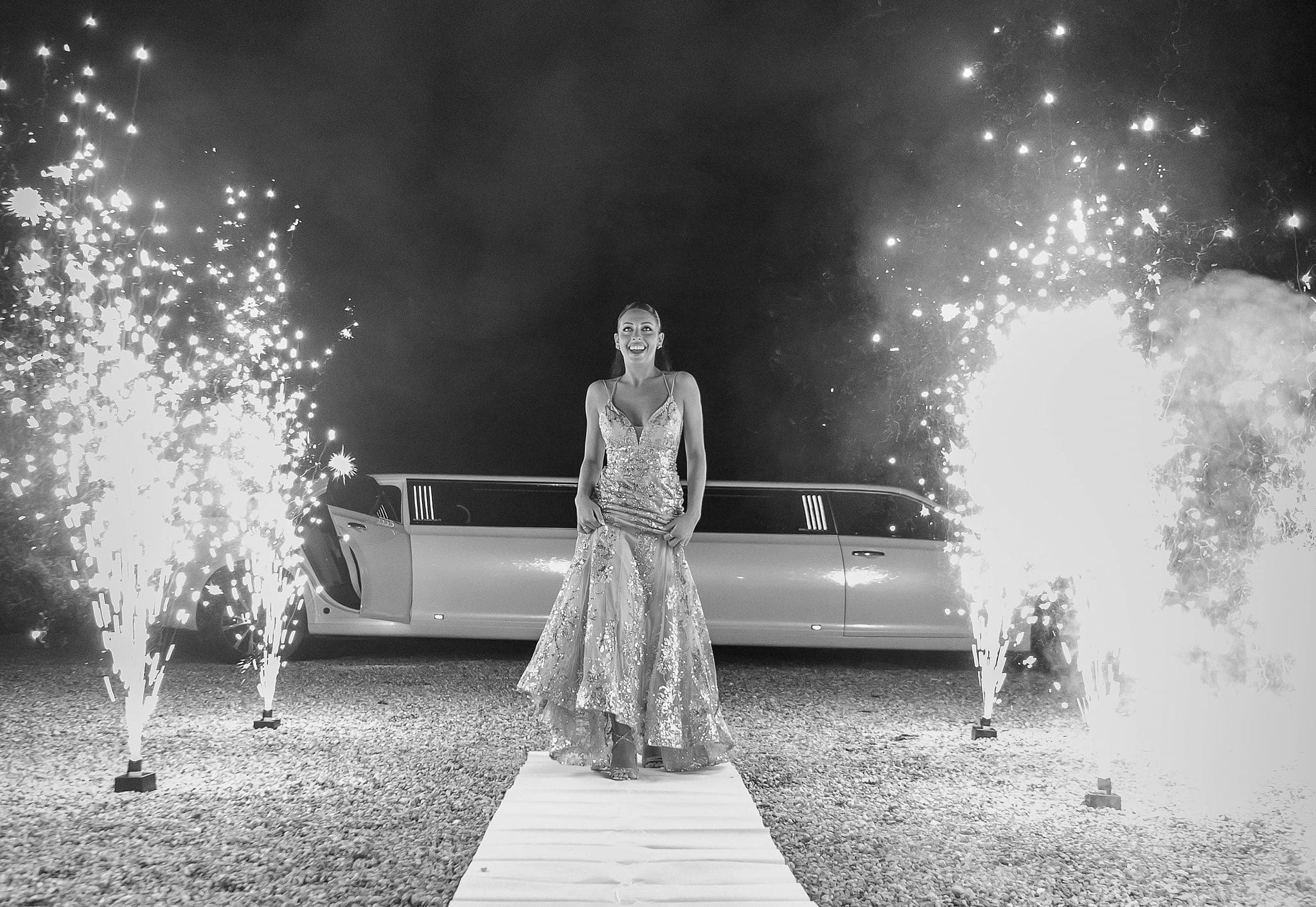 In bianco e nero, la festeggiata in abito lungo cammina su un tappeto bianco tra due fontane di scintille, con la limousine alle spalle