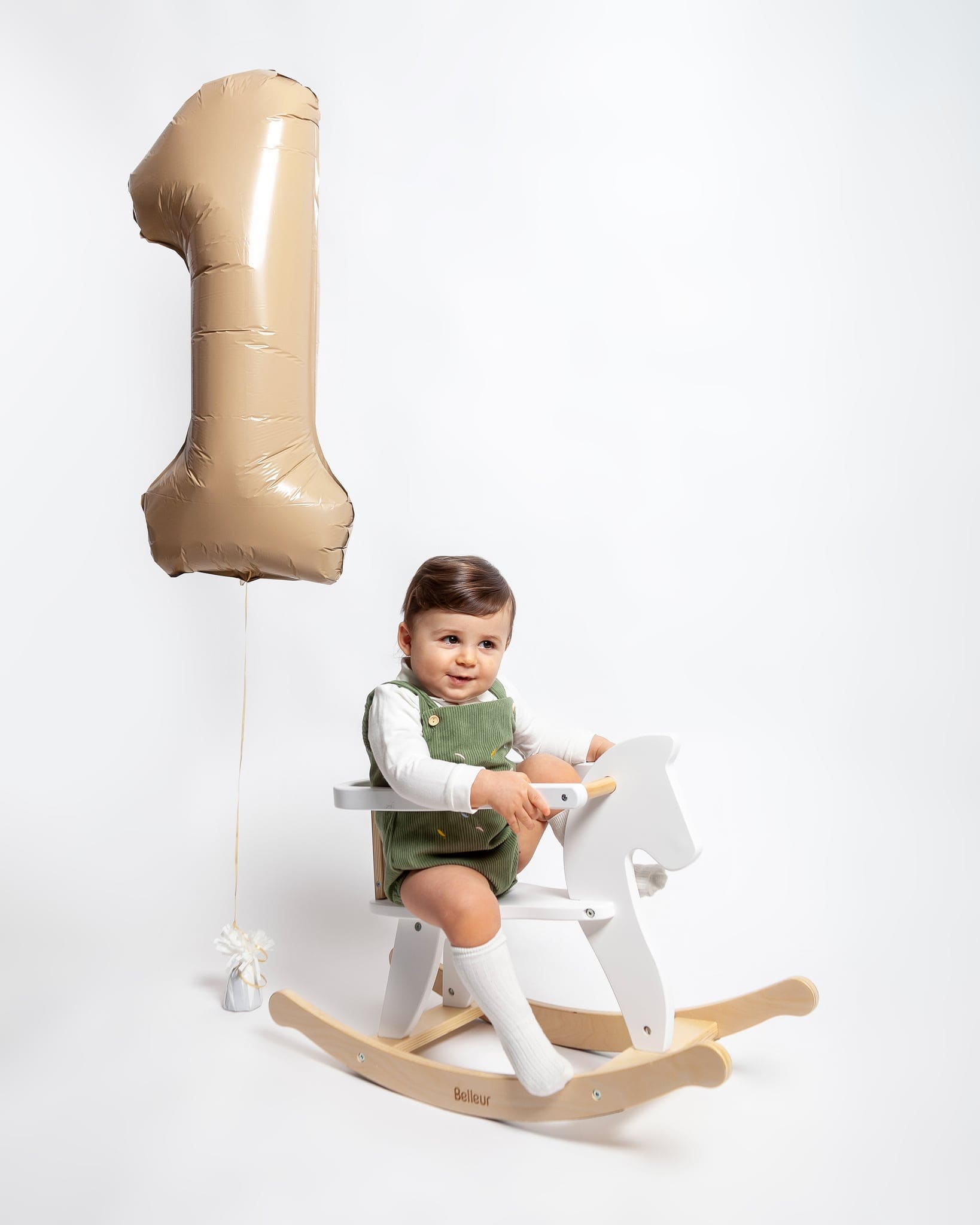 Il bimbo in salopette verde e calzini bianchi siede su un cavallo a dondolo, con un palloncino beige a forma di uno