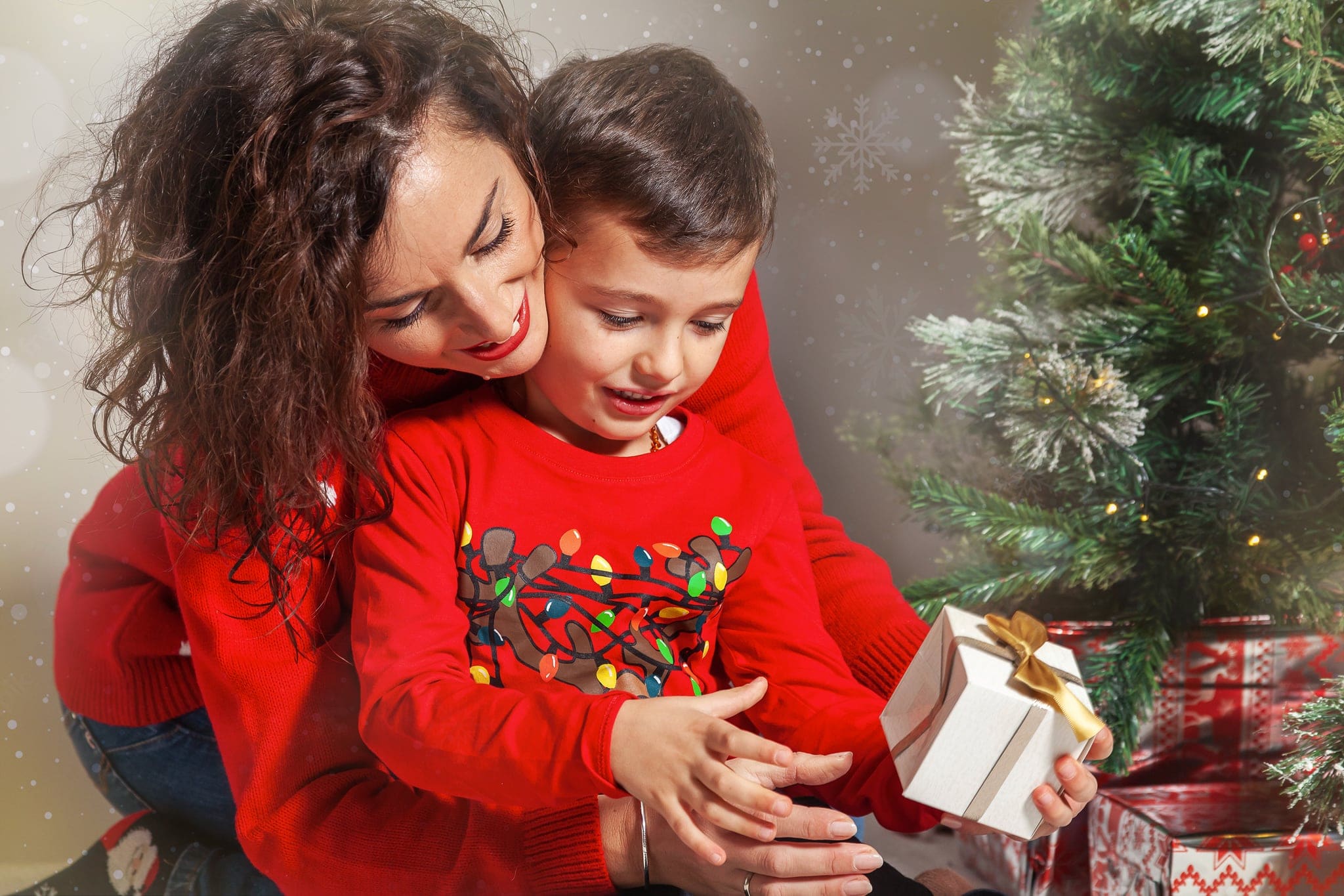 Mamma abbraccia il figlio in maglione rosso con luci colorate mentre apre un regalo con fiocco dorato, vicino all'albero