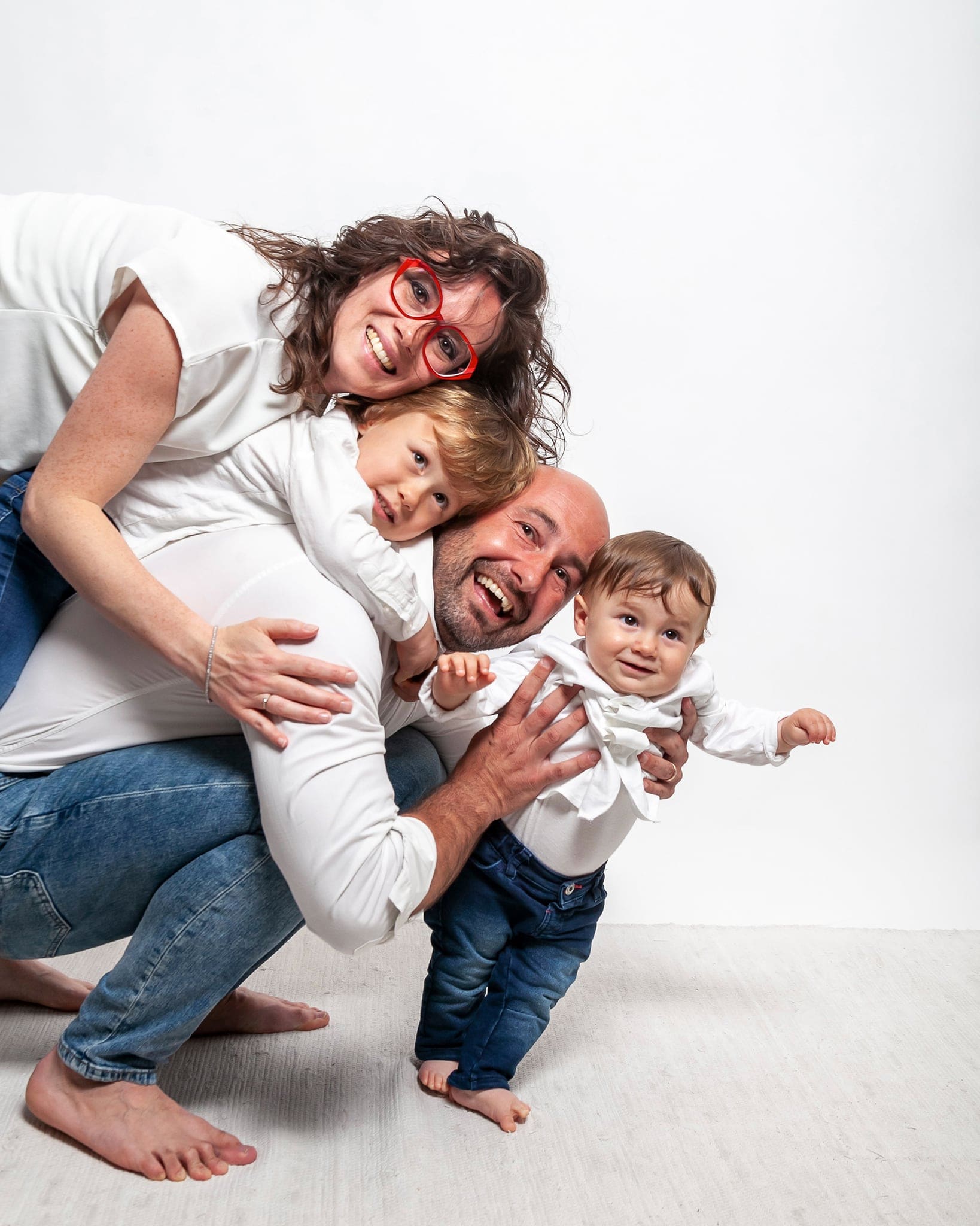 Famiglia che ride in studio su fondo bianco: mamma con occhiali rossi, papà e due bimbi piccoli sorridenti