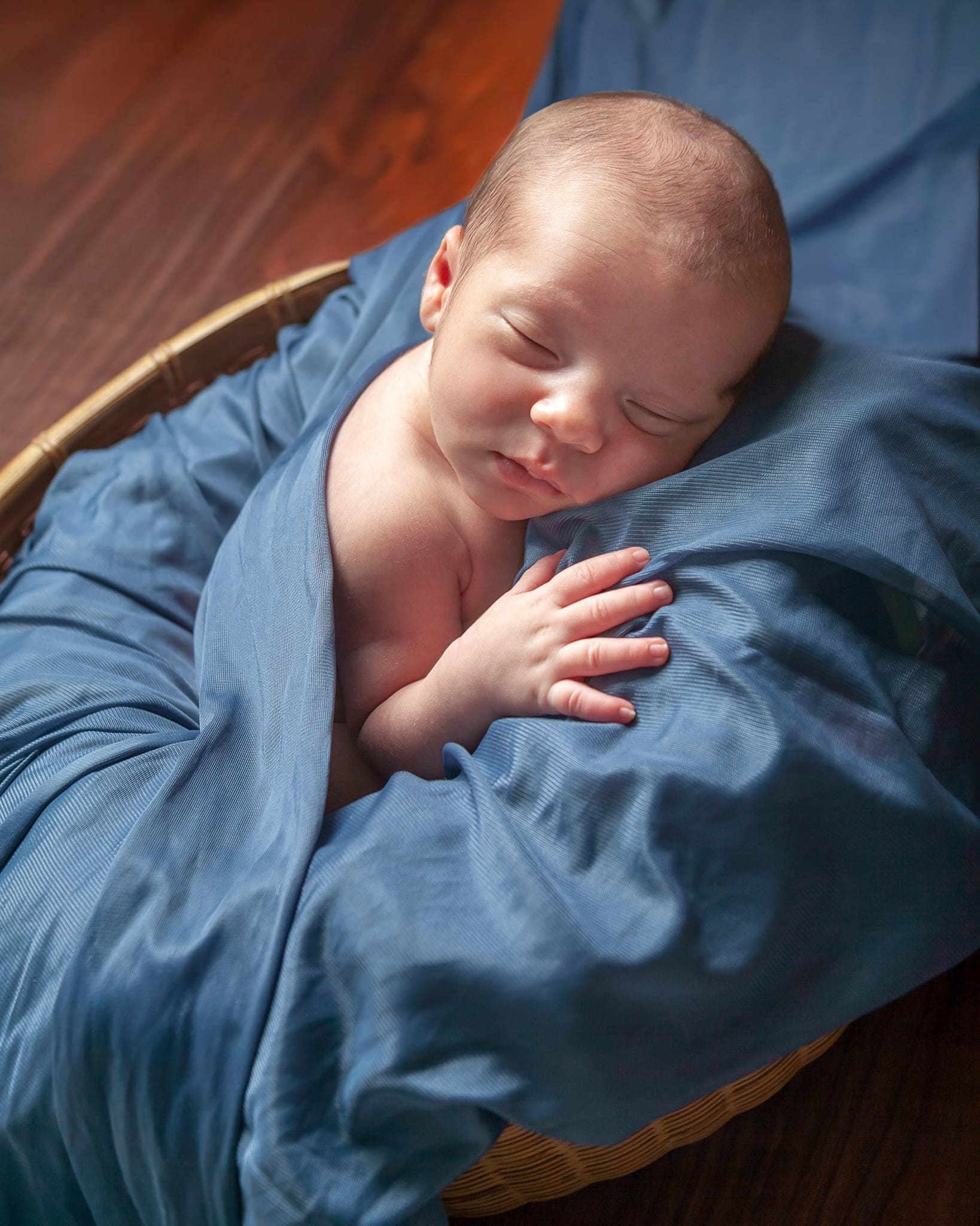 Neonato che dorme avvolto in un telo di seta blu, con una manina appoggiata sul tessuto, in un cesto su fondo di legno scuro.