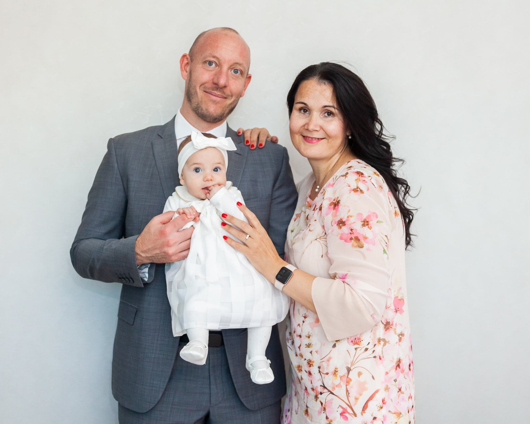 Ritratto di papà e mamma sorridenti che tengono in braccio la piccola con l'abito bianco del battesimo, su sfondo chiaro