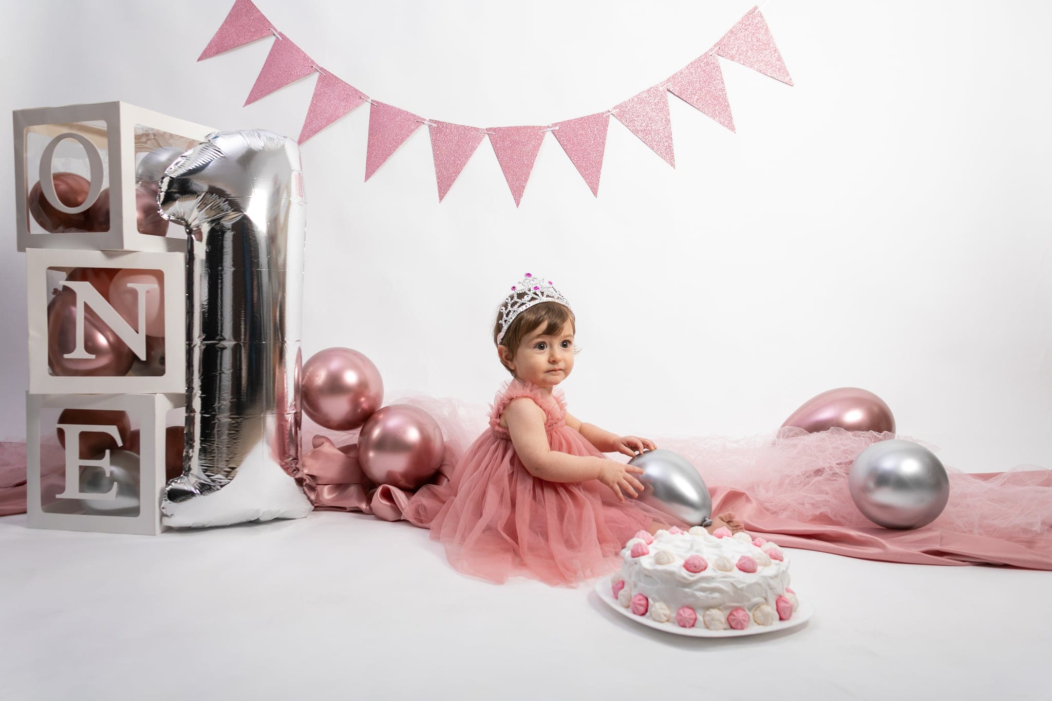La piccola con una coroncina sul capo siede tra palloncini argentati e rosa, accanto ai cubi bianchi con la scritta ONE e alla torta