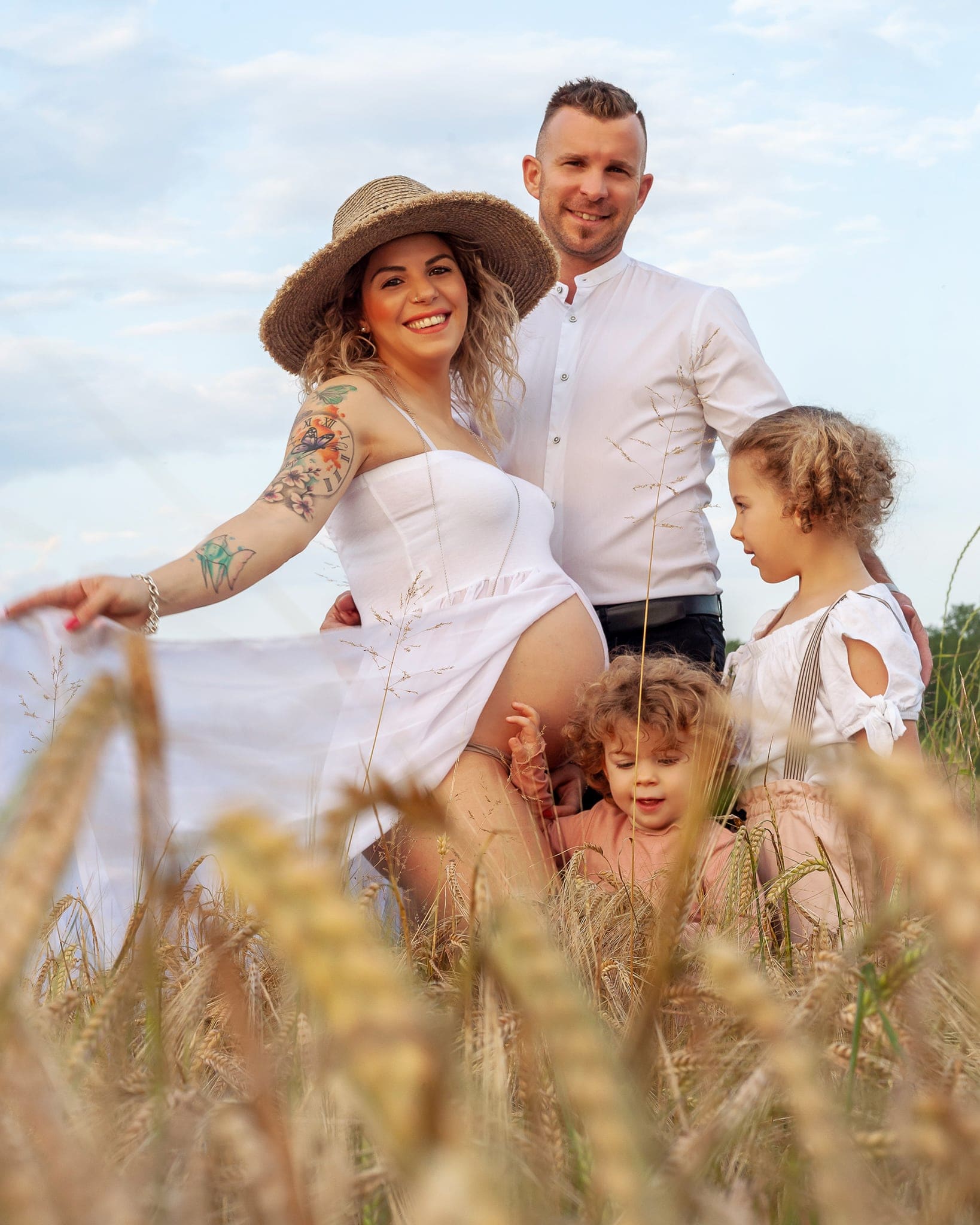 Futura mamma con cappello di paglia e abito bianco con papà e due bambini tra le spighe di grano, shooting di gravidanza all'aperto.