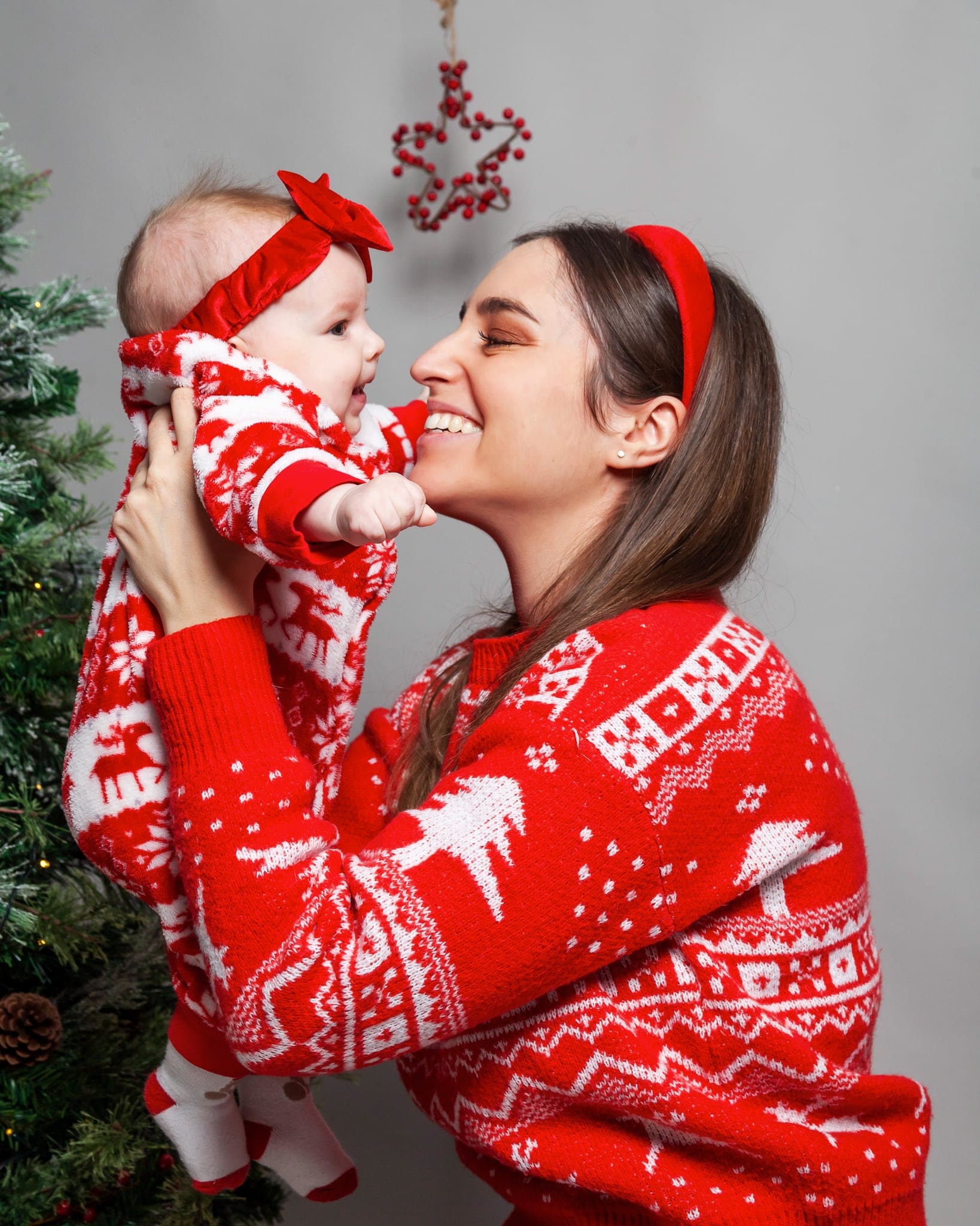 Mamma con cerchietto rosso solleva la neonata in tutina natalizia e le sorride, entrambe in rosso, davanti all'albero