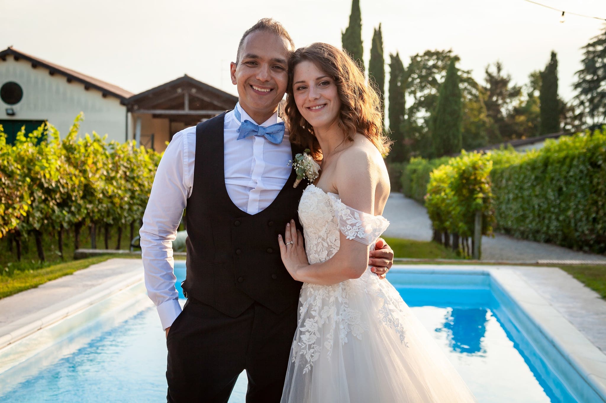 Sposi sorridenti abbracciati accanto a una piscina, con vigneti e cipressi sullo sfondo, matrimonio sul Collio