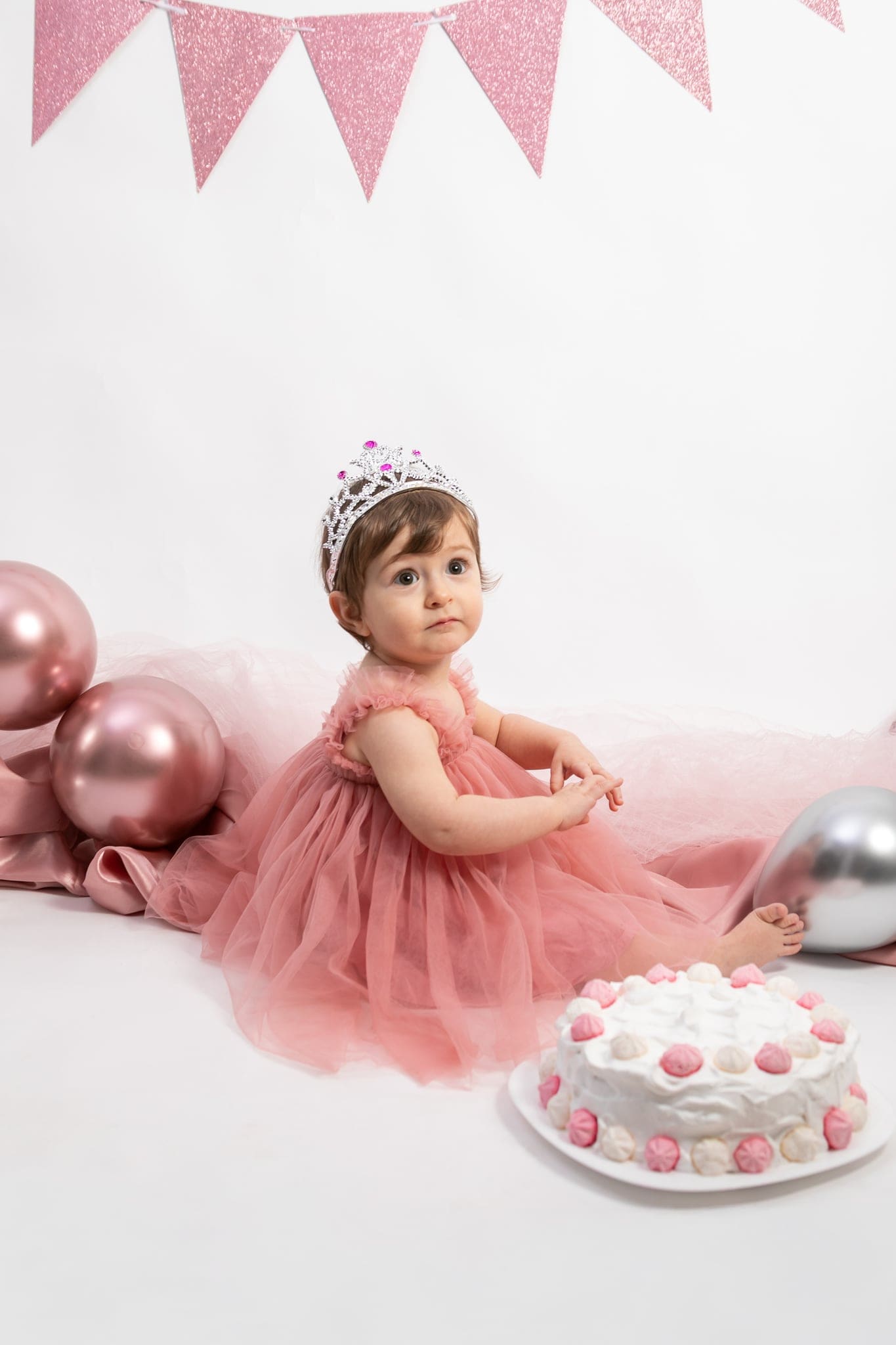 Seduta con la coroncina e il vestito di tulle rosa, la piccola guarda in camera accanto alla torta bianca con meringhe rosa