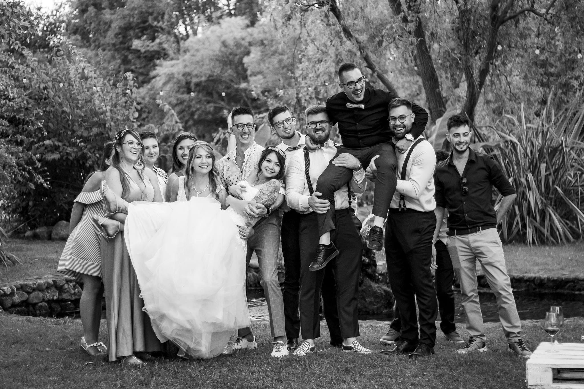Gruppo di amici ridono in bianco e nero mentre sollevano lo sposo in braccio alla sposa, festa di matrimonio a Rivignano Teor
