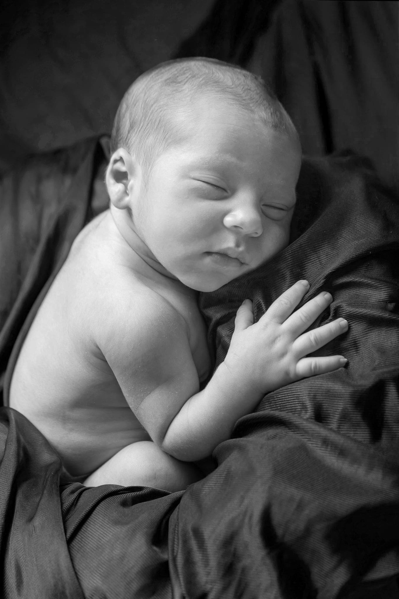 Neonato addormentato con la manina sul petto, in bianco e nero, su un tessuto lucido scuro in uno scatto morbido e contrastato.