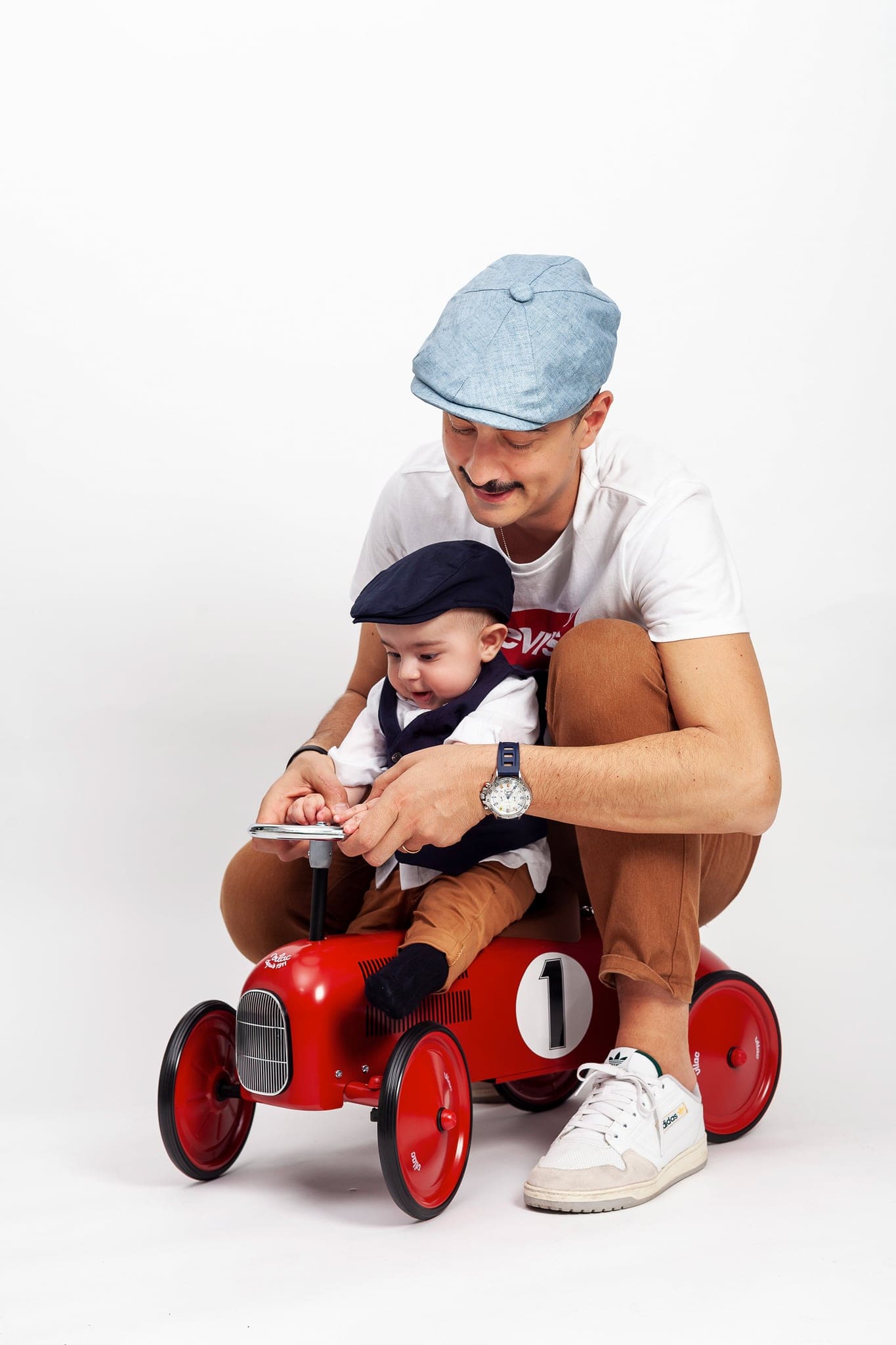 Papà con cappello azzurro e bimbo piccolo seduti su una macchinina rossa d'epoca con il numero uno, sfondo bianco