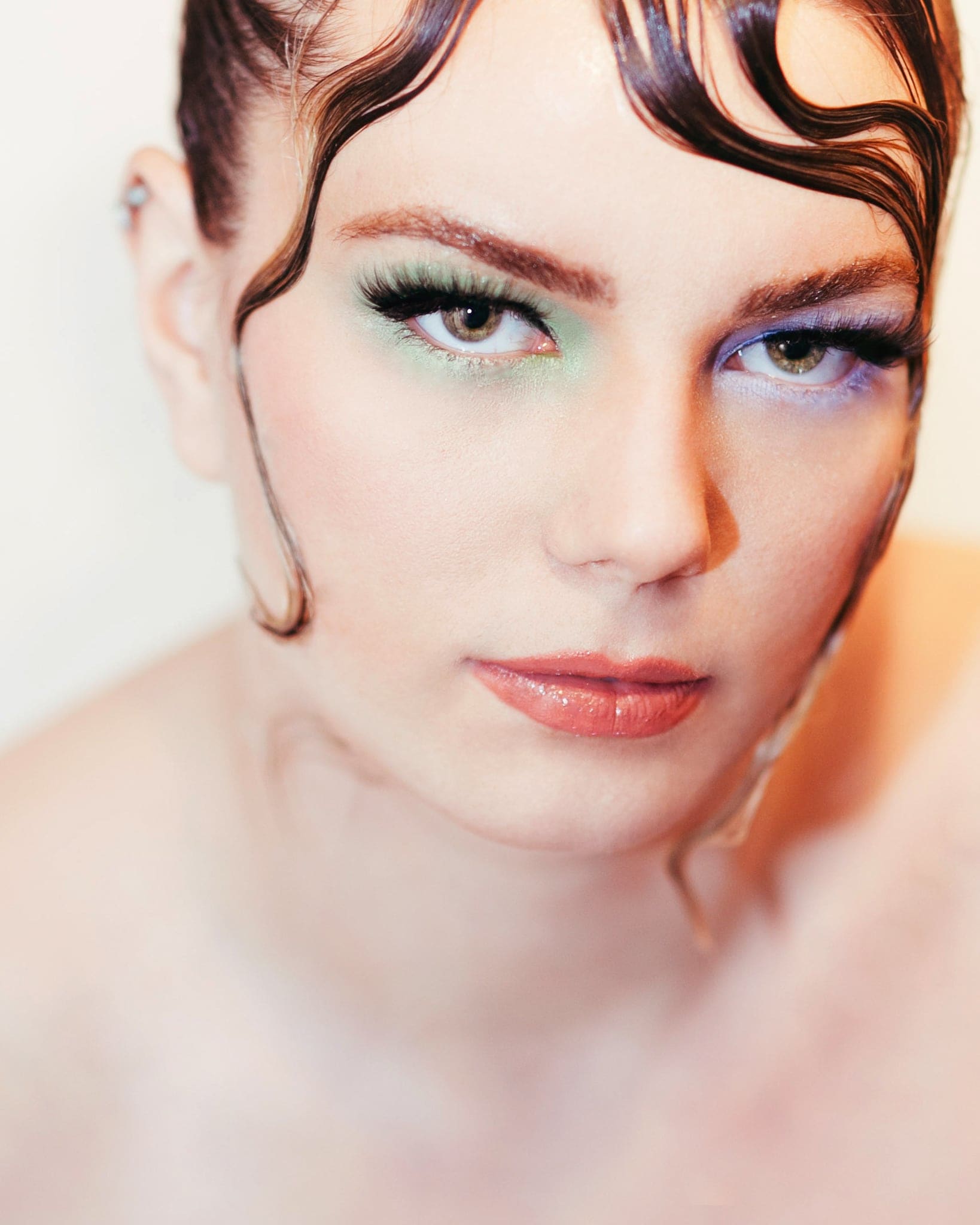Primo piano di una donna con trucco artistico verde e viola sugli occhi e ciocche di capelli ondulate sul viso, luce morbida e chiara