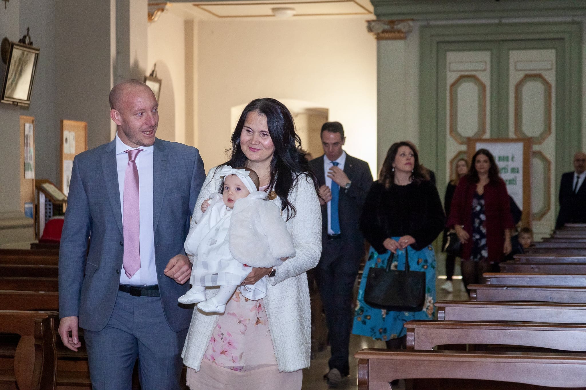 I genitori entrano in chiesa tra le panche con la piccola in abito bianco in braccio, seguiti dagli invitati