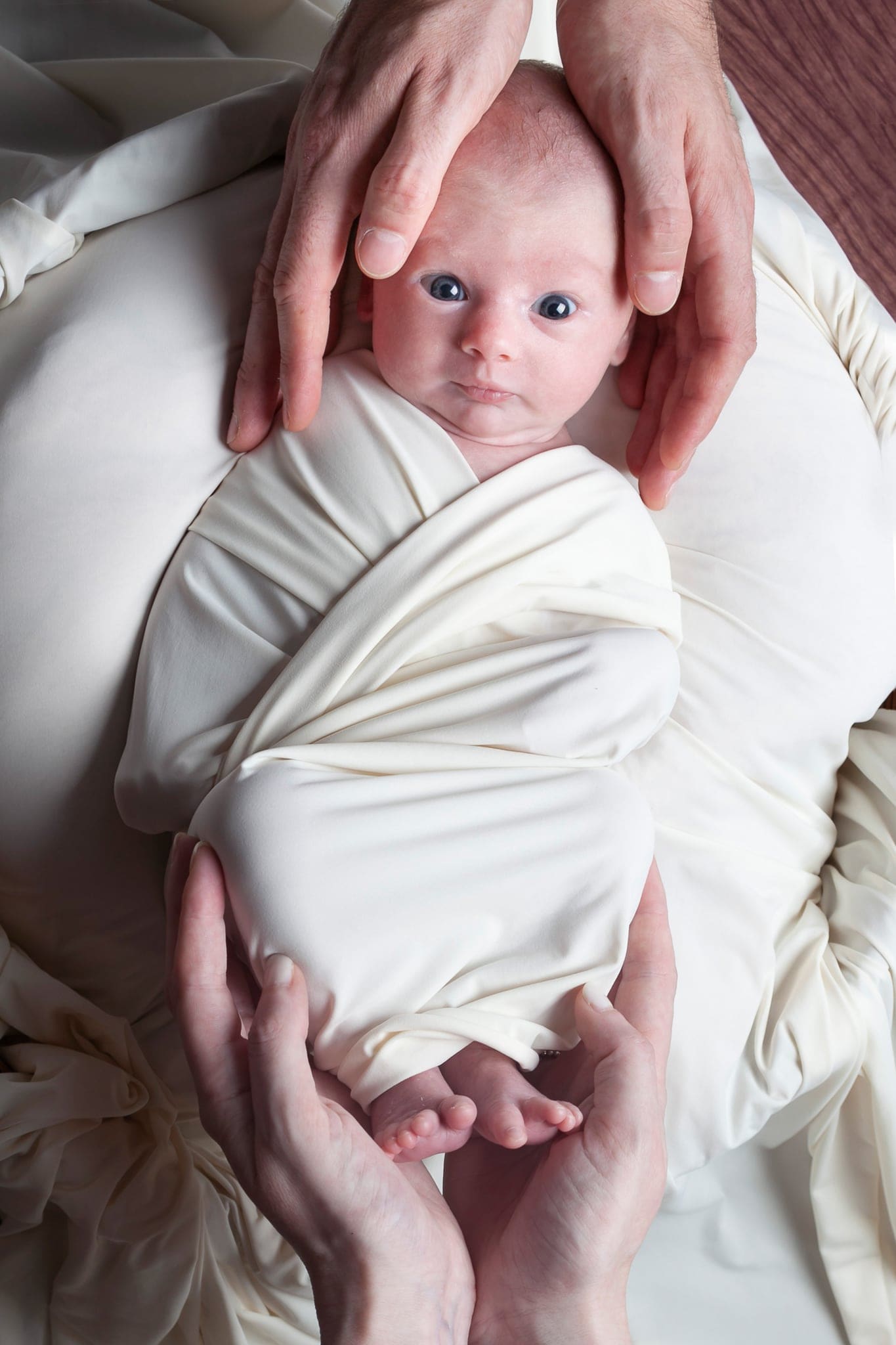 Neonato avvolto in fasce color panna con il viso sveglio, tenuto delicatamente per la testa e i piedini da due paia di mani.