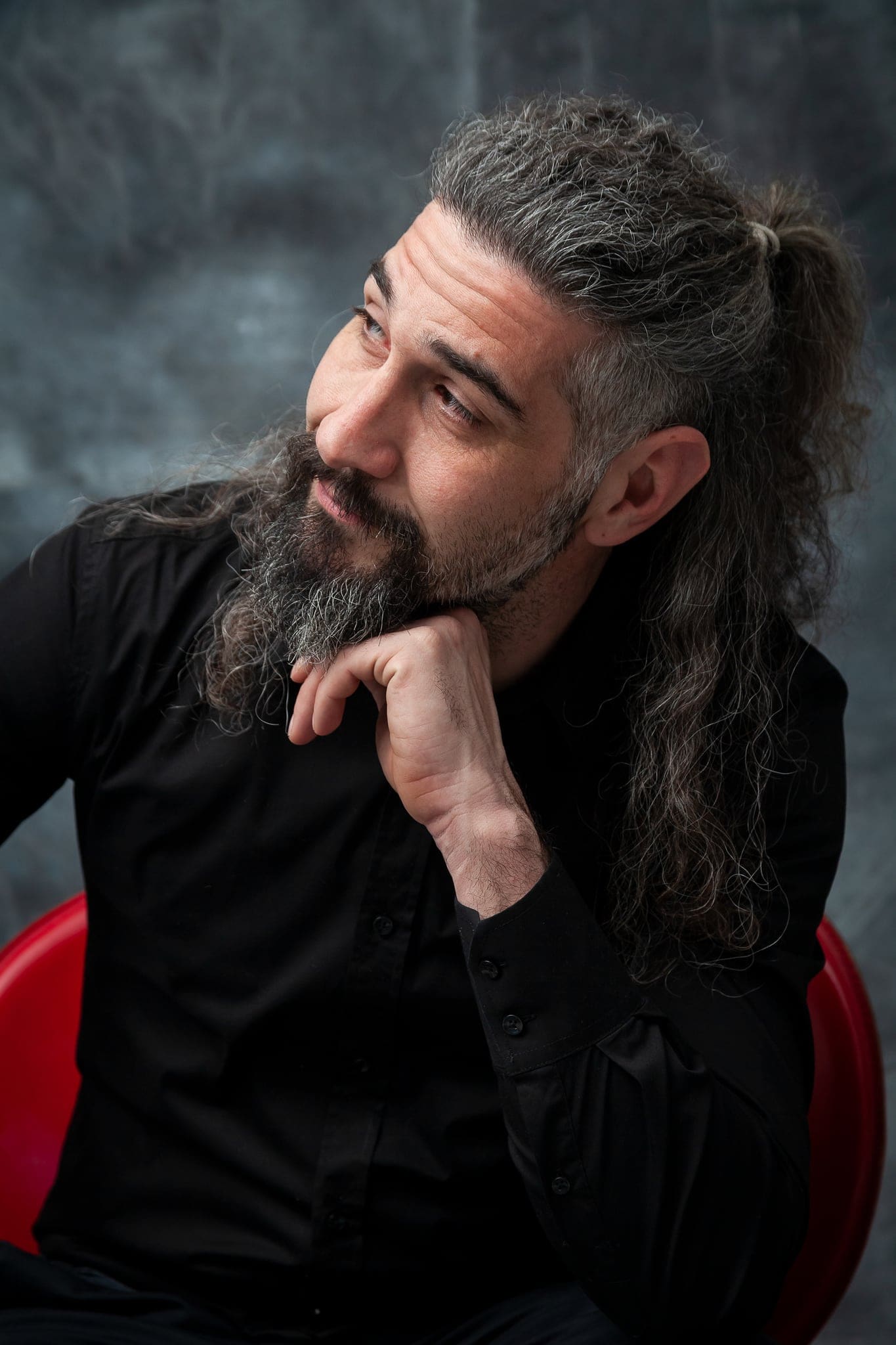 Uomo con lunghi capelli grigi raccolti e barba in camicia nera, mano al mento e sguardo di lato, seduto su una sedia rossa