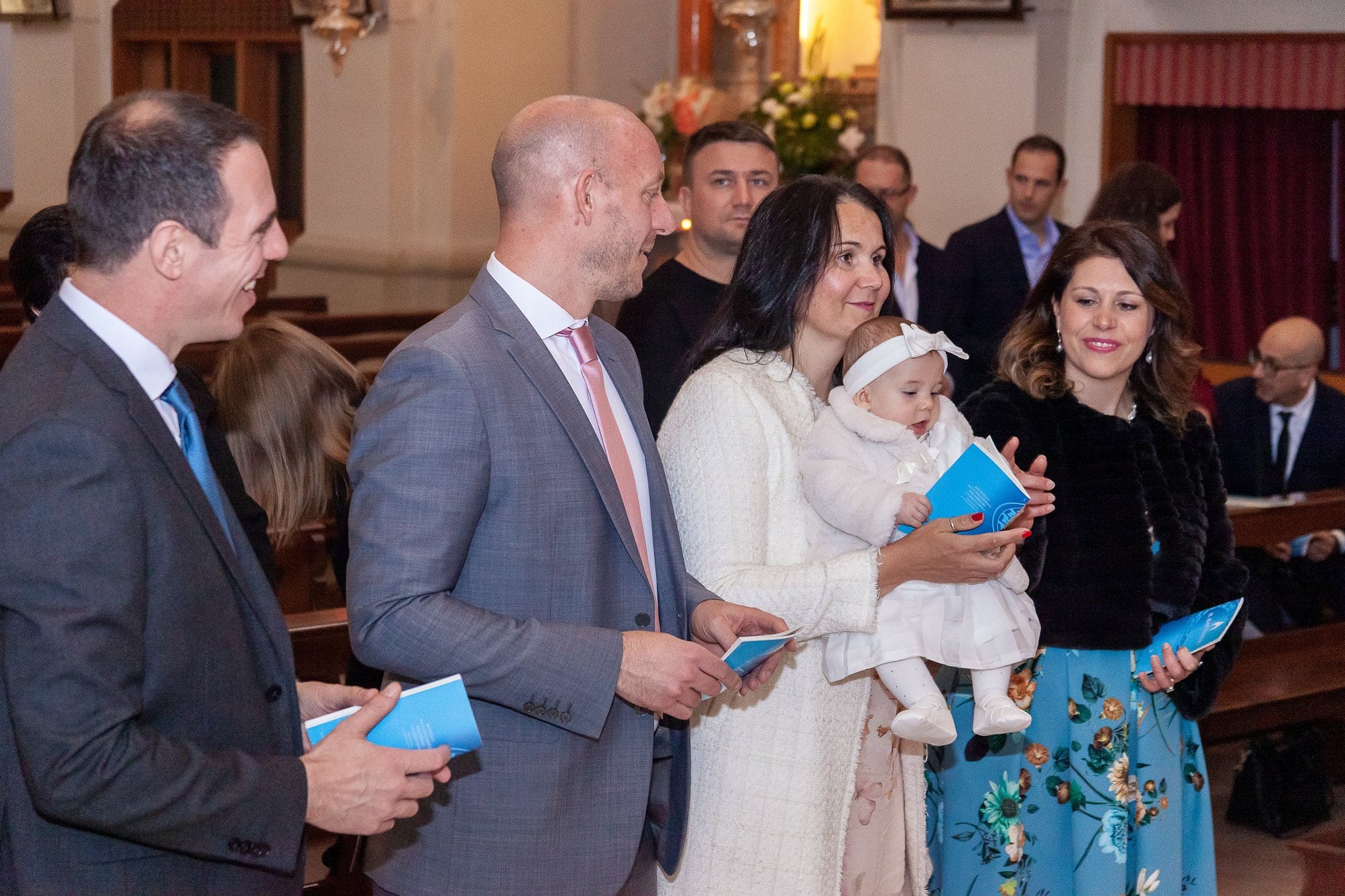 Durante la celebrazione, due uomini e due donne con i libretti azzurri guardano sorridendo la piccola in braccio alla mamma