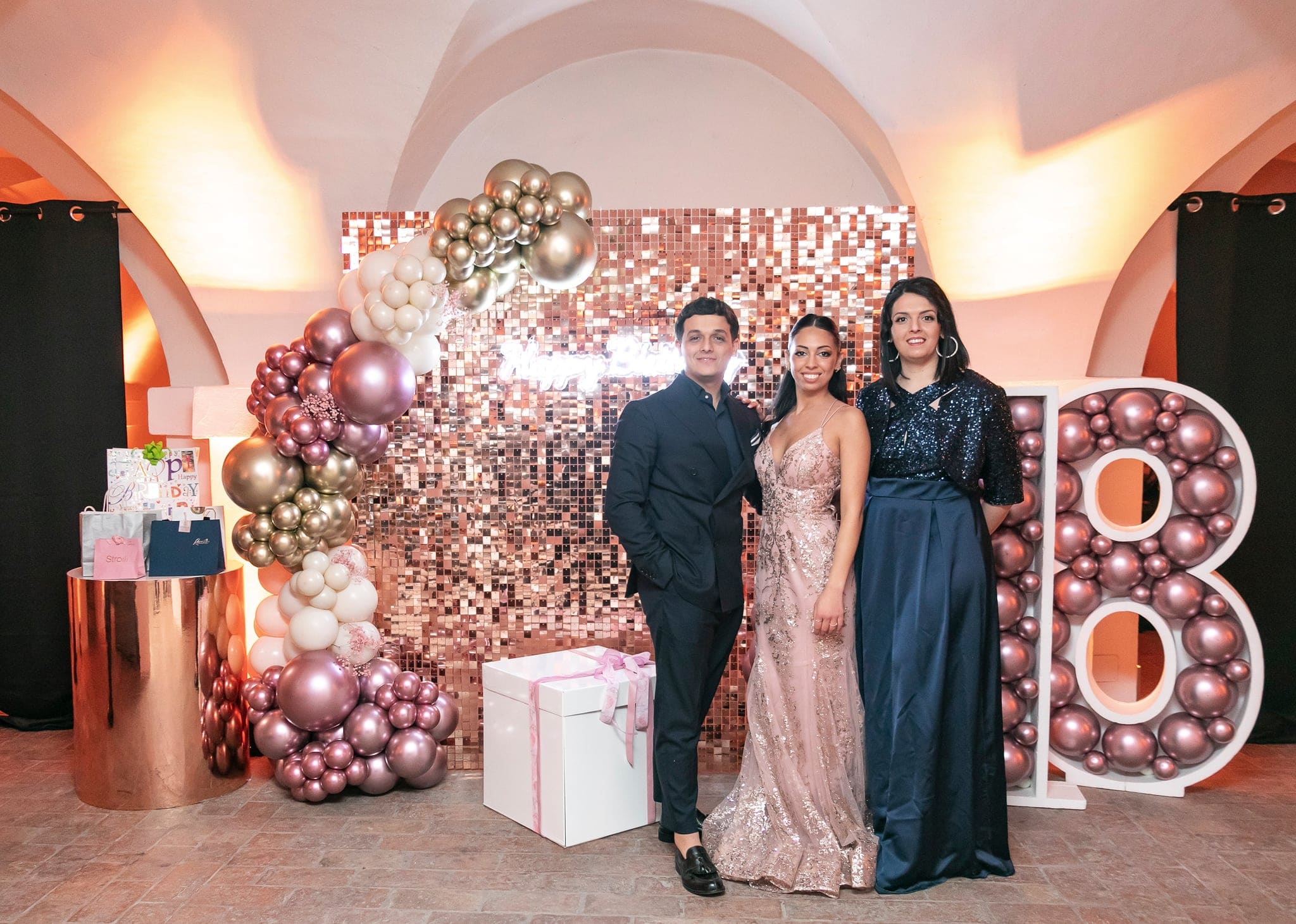 La festeggiata con un ragazzo e una donna in abito blu in posa davanti alla parete di paillettes e al grande numero 18 di palloncini