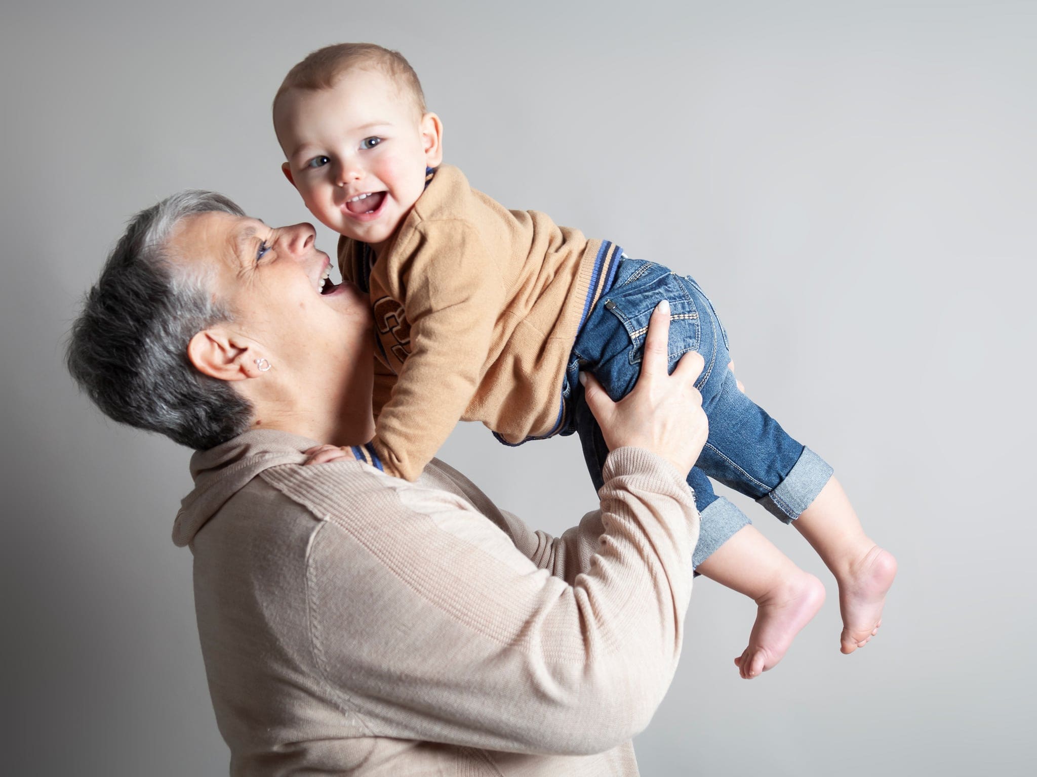 Nonno sorridente solleva in alto un bimbo piccolo che ride, ritratto in studio su fondo grigio