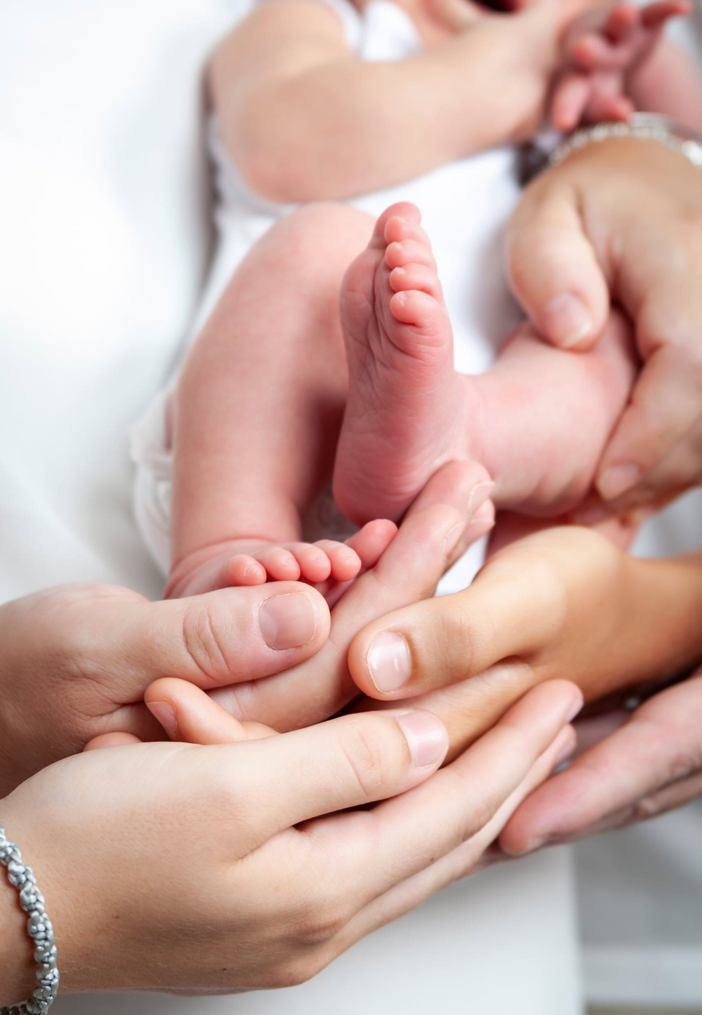 Piedini di un neonato stretti e accarezzati da diverse mani di adulti, su fondo chiaro luminoso, in un servizio newborn in studio.