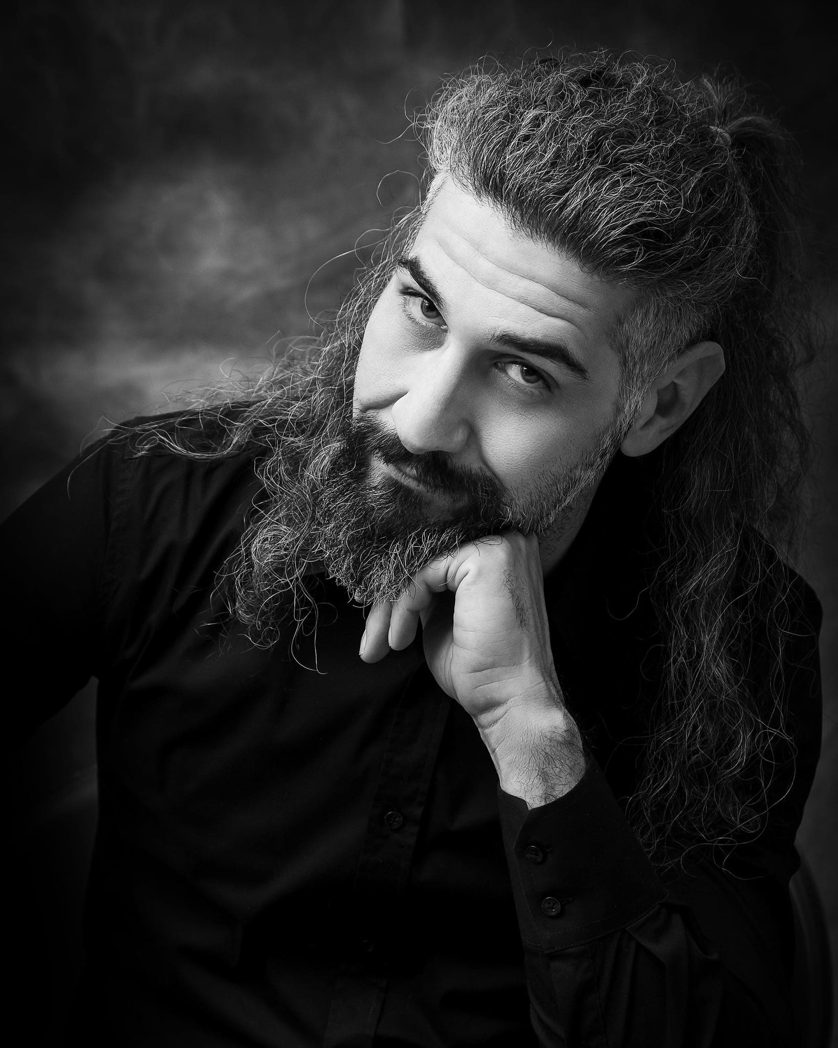 Ritratto in bianco e nero di un uomo con lunghi capelli e barba folta, mano al mento, in camicia scura su fondo sfumato
