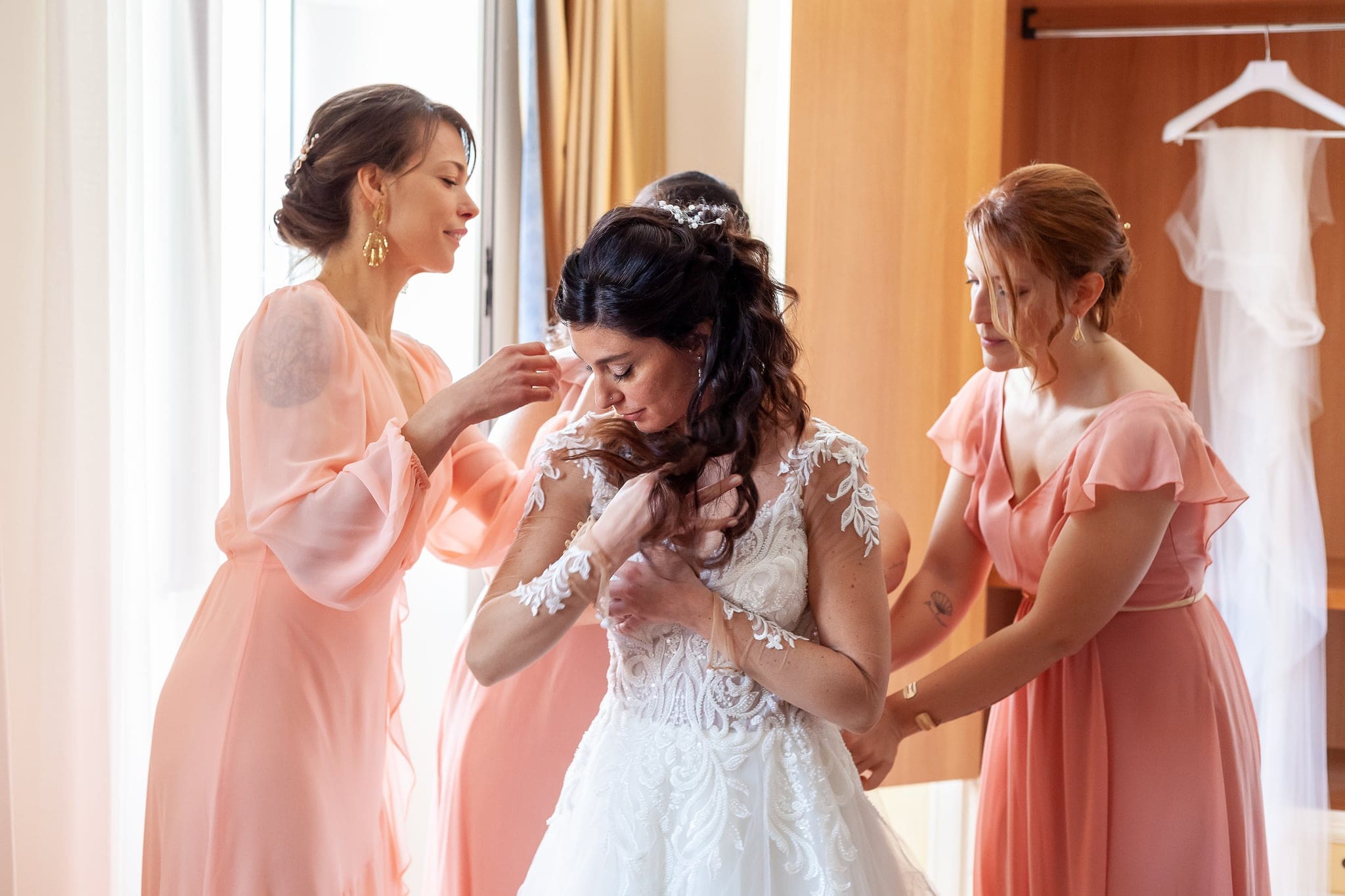 Due damigelle in abito rosa aiutano la sposa ad allacciare l'abito vicino alla finestra durante la vestizione a Trieste
