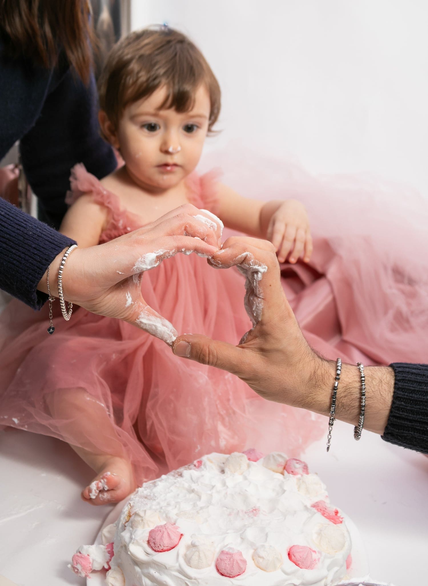 Le mani sporche di panna di mamma e papà formano un cuore davanti alla piccola in tulle rosa, vicino alla torta