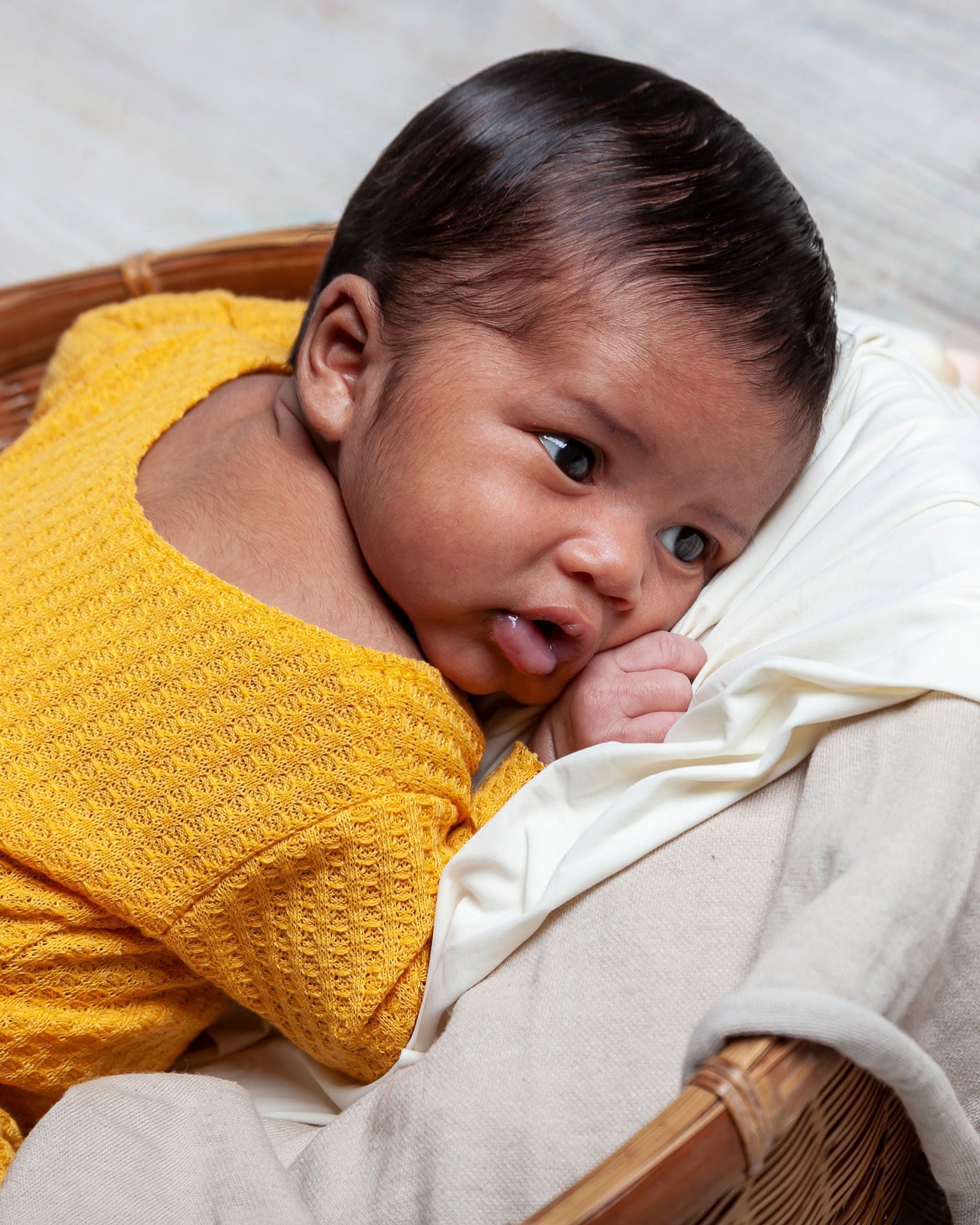 Primo piano di un neonato con tutina gialla che appoggia il viso su una coperta, con lo sguardo curioso e la lingua appena fuori.