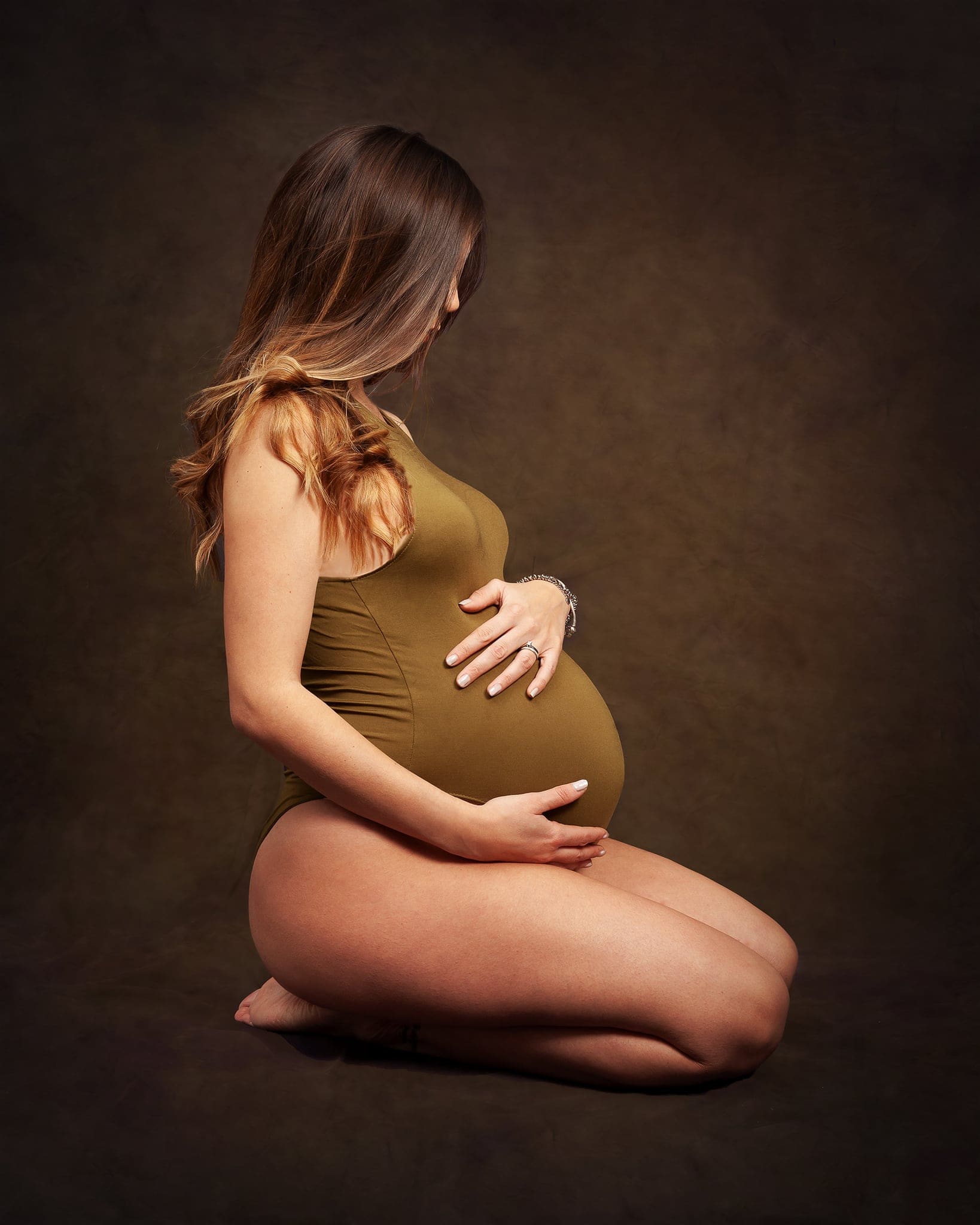 Donna incinta inginocchiata di profilo con body verde oliva, che accarezza il pancione, su sfondo marrone scuro.