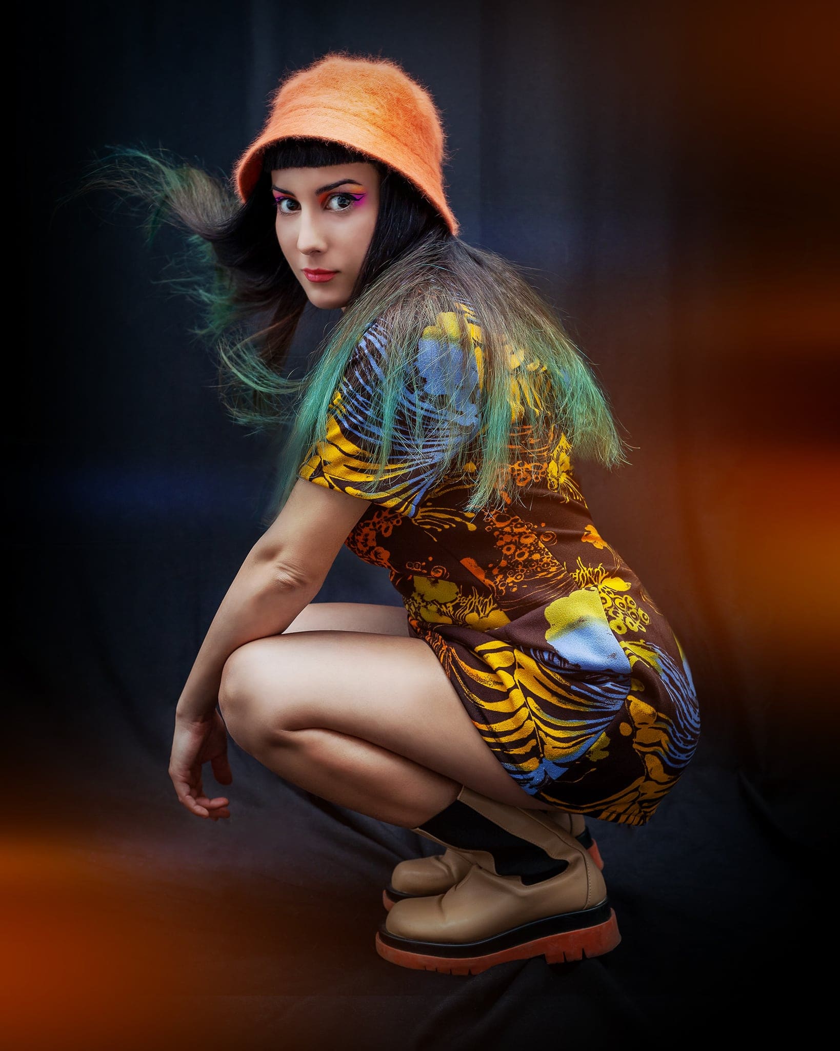Ragazza con cappello arancione e capelli con sfumature verdi, in abito colorato e scarponcini, accovacciata su fondo scuro con luci calde
