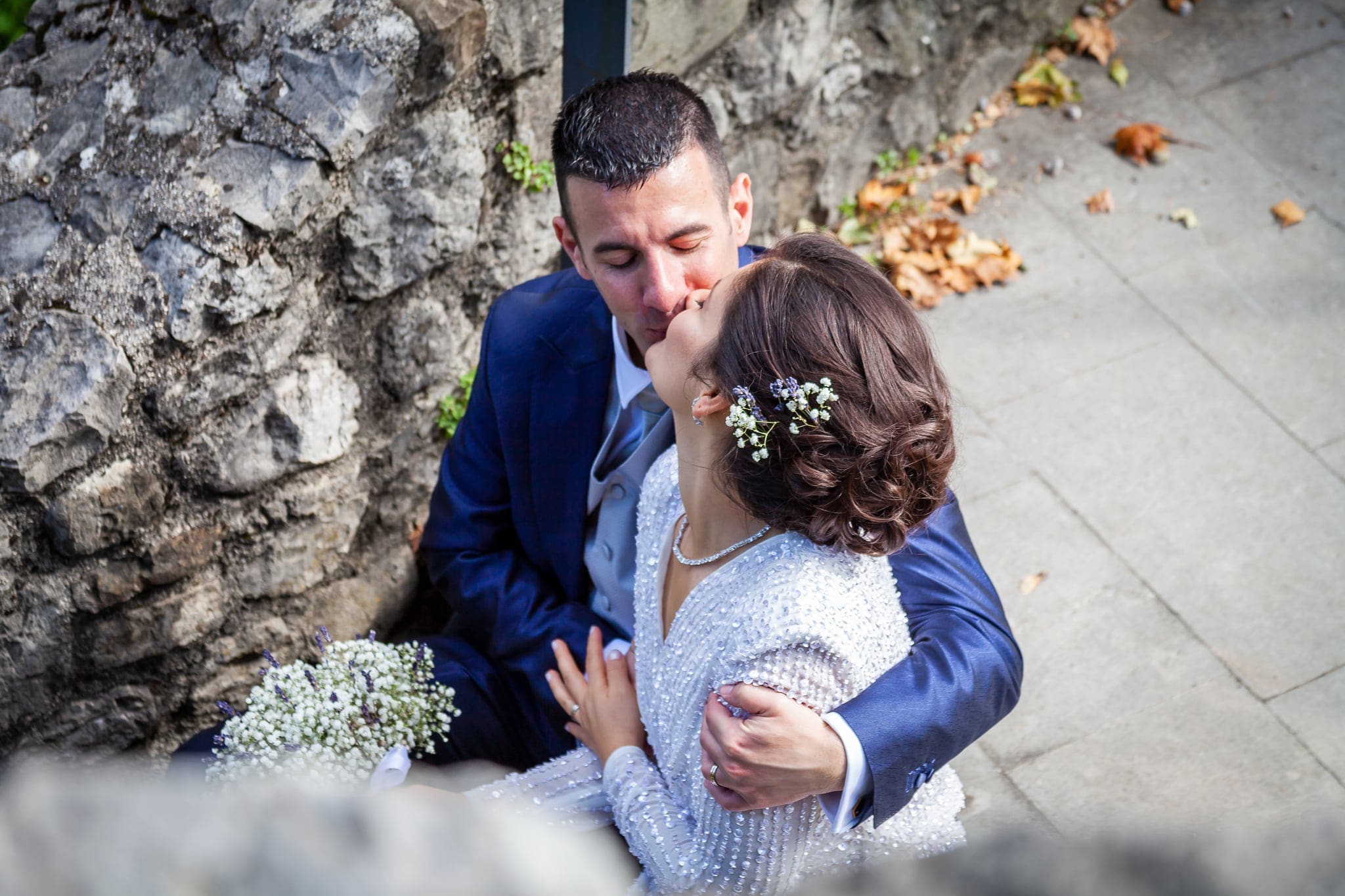 Sposo e sposa seduti accanto a un muro di pietra si scambiano un bacio, con il bouquet bianco, matrimonio a Cividale del Friuli