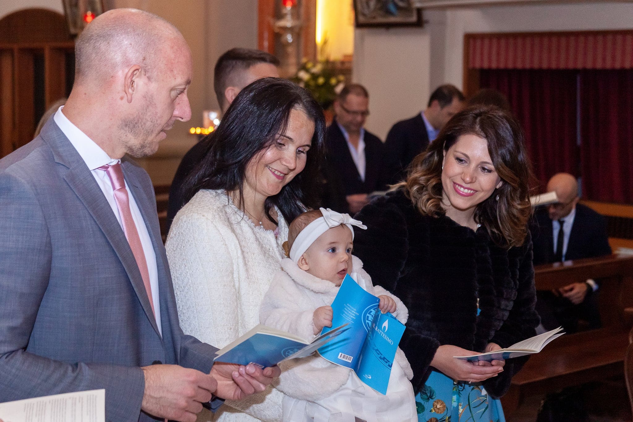 Primo piano di mamma, papà e un'invitata che sorridono alla piccola con il fiocco bianco, durante la celebrazione