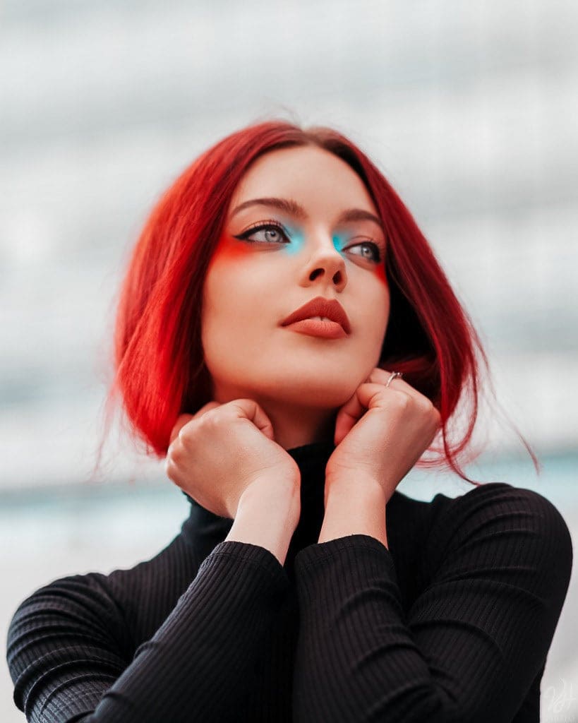 Ragazza dai capelli rossi e trucco azzurro, in dolcevita nero, con le mani sotto il mento e lo sguardo verso l'alto, sfondo chiaro sfocato