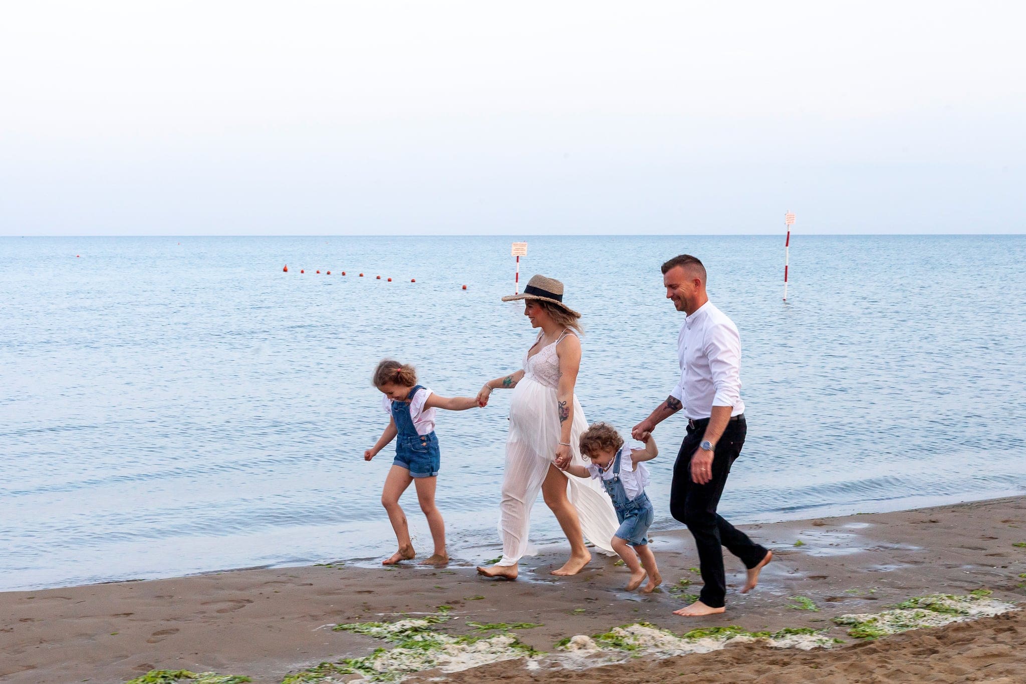 Famiglia in passeggiata sulla riva del mare al tramonto, mamma in attesa, papà e due bimbi che si tengono per mano