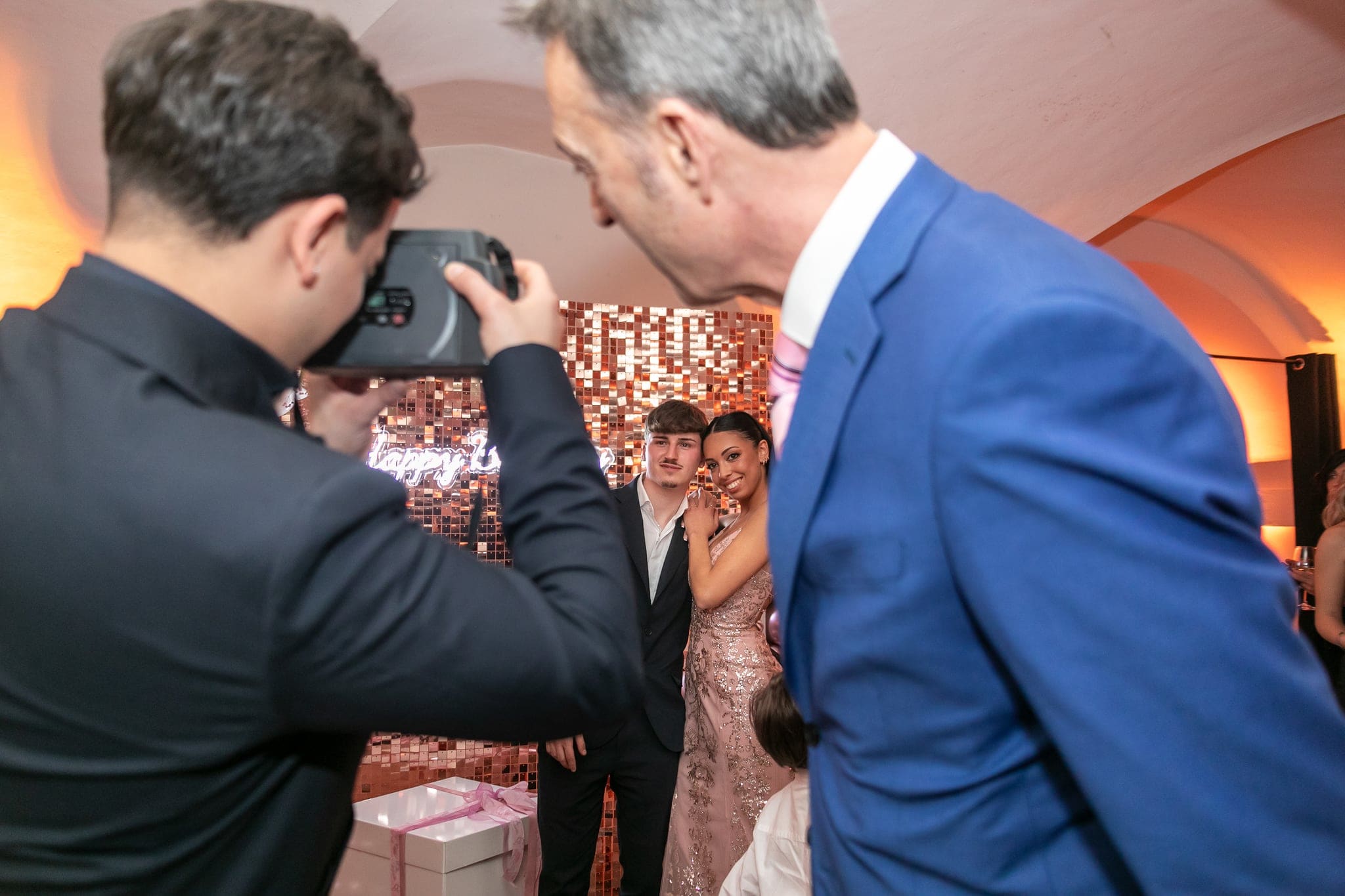 Due uomini fotografano la festeggiata e un ragazzo davanti alla parete di paillettes durante la serata dei 18 anni