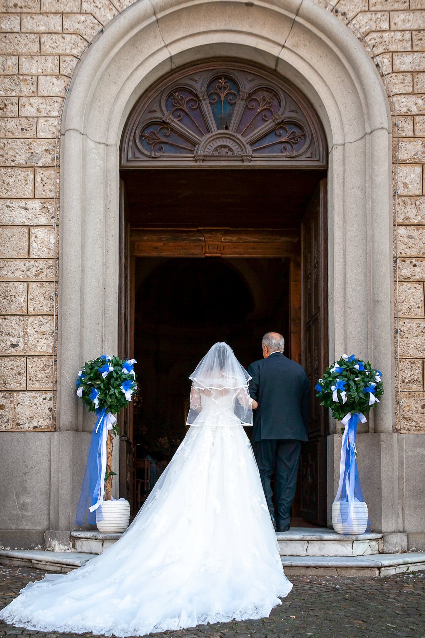 Sposa con lo strascico lungo entra in chiesa accompagnata da un uomo, tra due vasi di fiori con nastri blu, matrimonio in Friuli