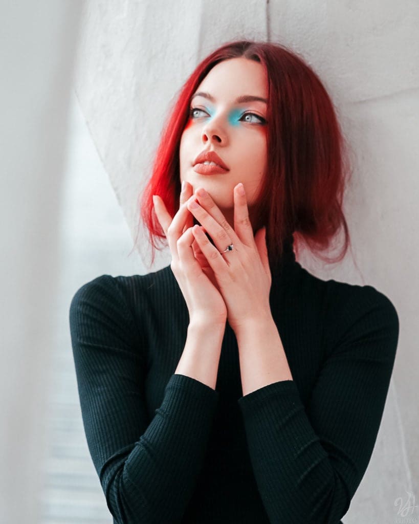 Ragazza dai capelli rossi e trucco azzurro in dolcevita nero, mani davanti al viso, appoggiata a un muro bianco e luminoso