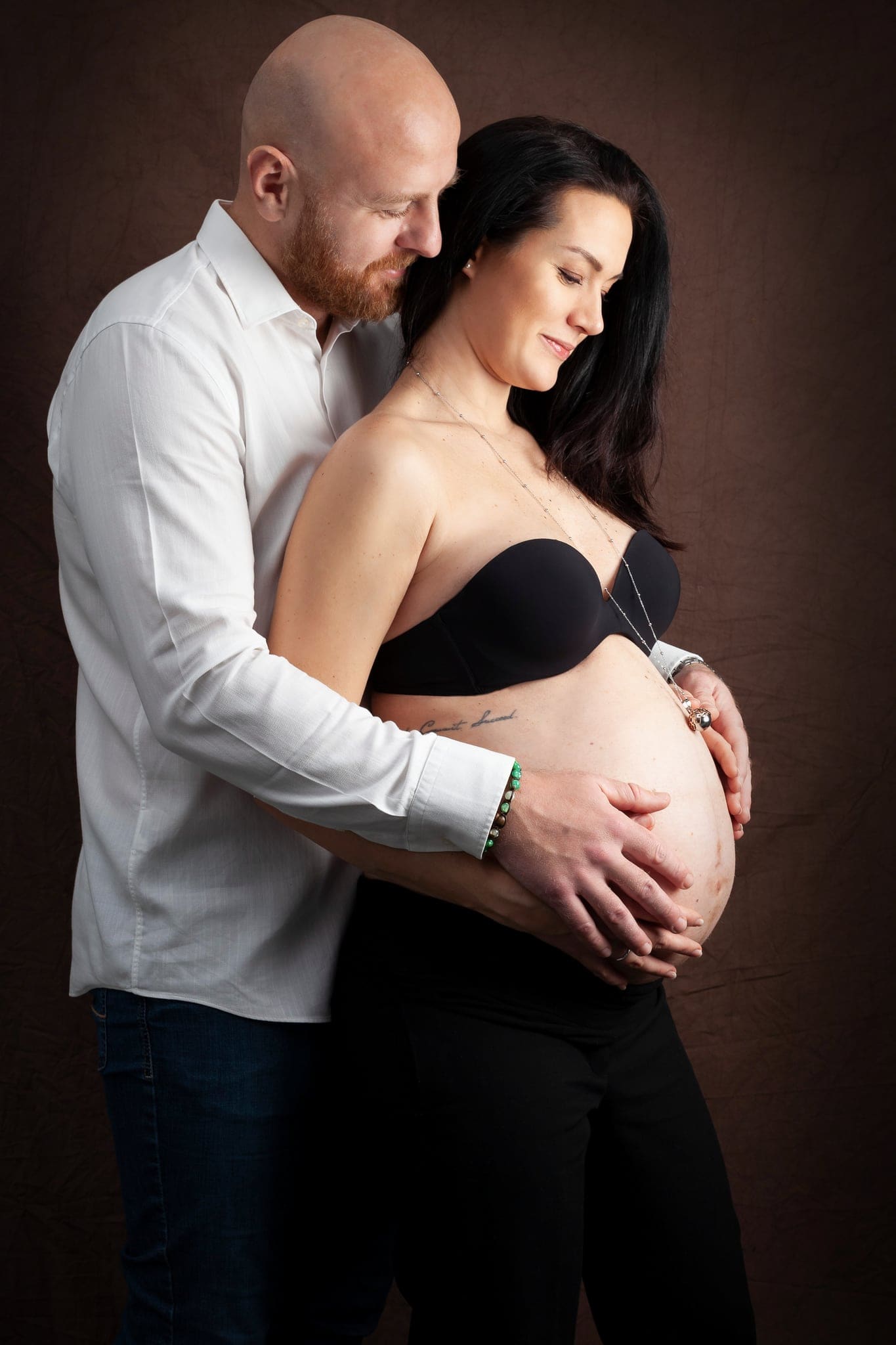 Papà con camicia bianca abbraccia da dietro la futura mamma in reggiseno e pantaloni neri, con le mani sul pancione, su fondo marrone.
