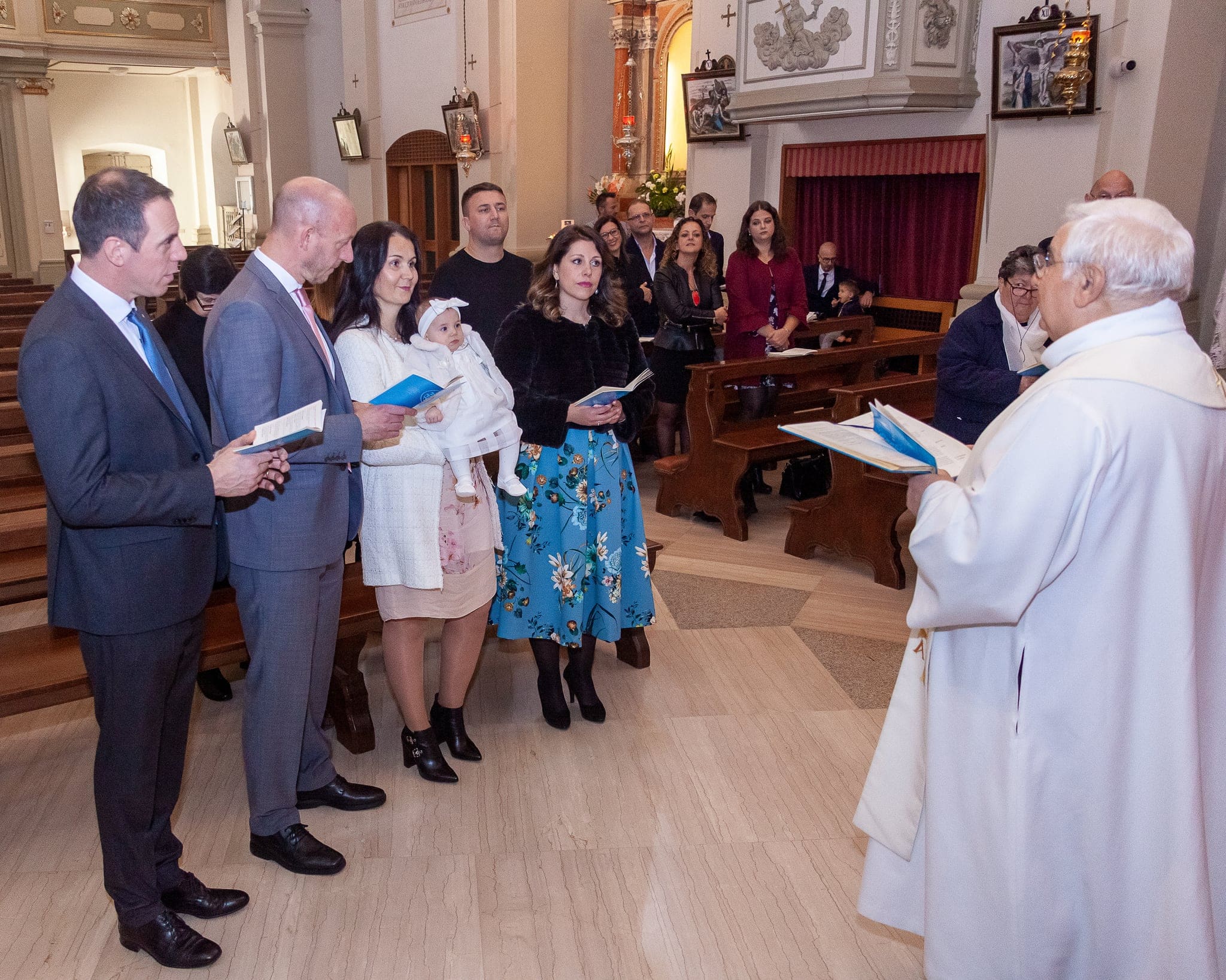 Il sacerdote in camice bianco davanti alla famiglia schierata in chiesa, con la bimba in braccio alla mamma e i libretti in mano