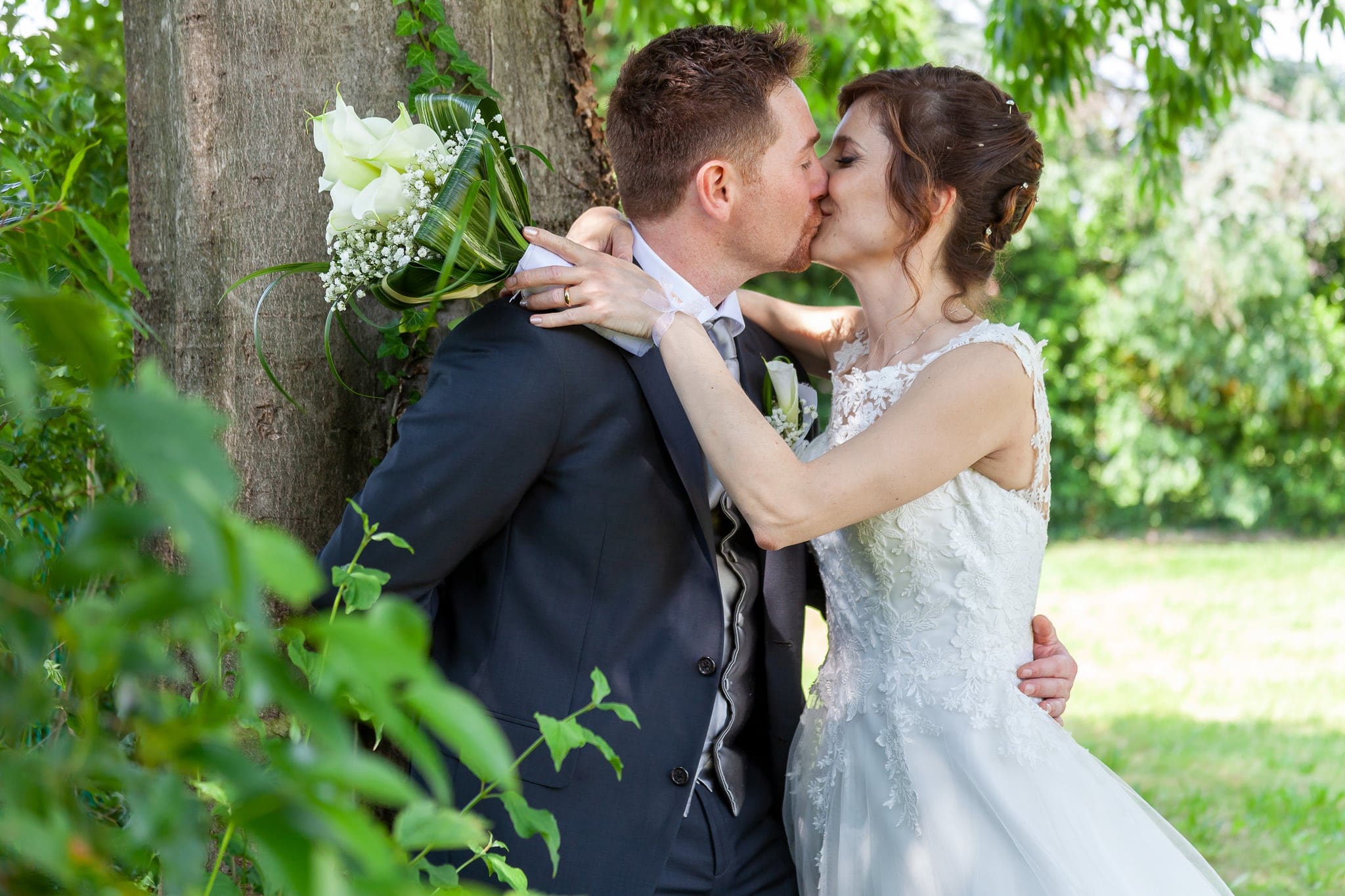 Sposi che si baciano appoggiati a un albero nel verde, con una rosa bianca nel bouquet, matrimonio in Friuli Venezia Giulia
