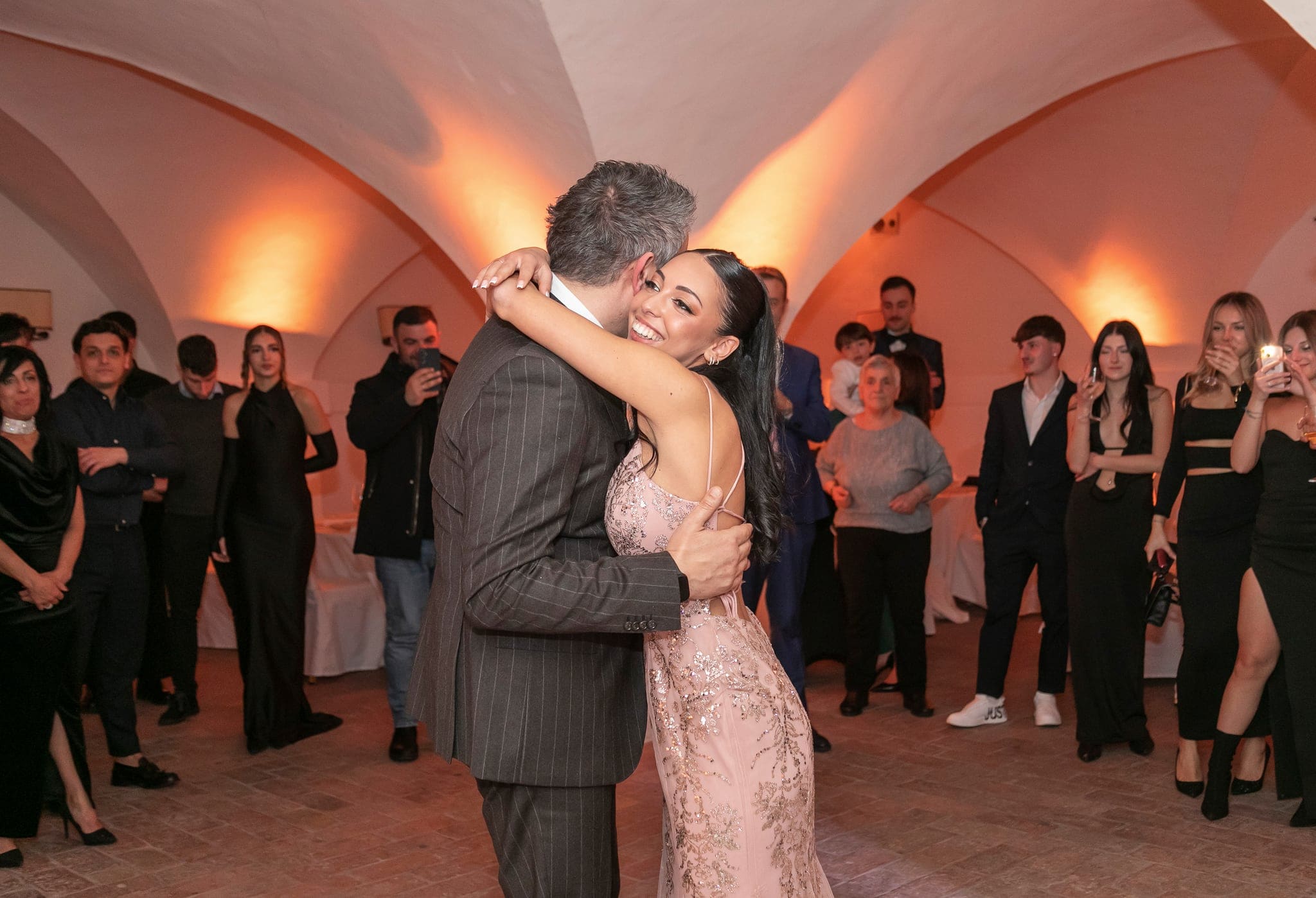 La festeggiata in abito rosa abbraccia un uomo in completo gessato al centro della sala, mentre gli invitati li guardano