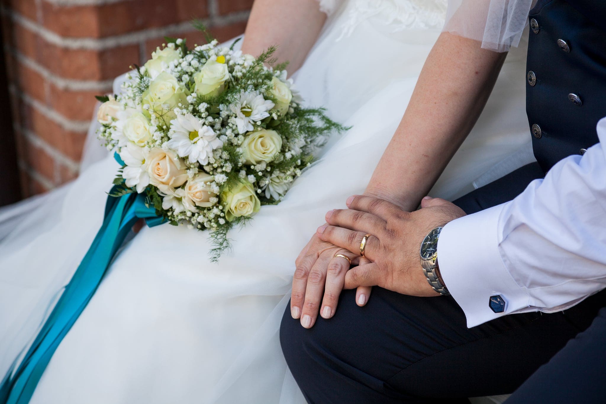 Primo piano del bouquet bianco con nastro turchese e delle mani degli sposi con le fedi, matrimonio a Pagnacco