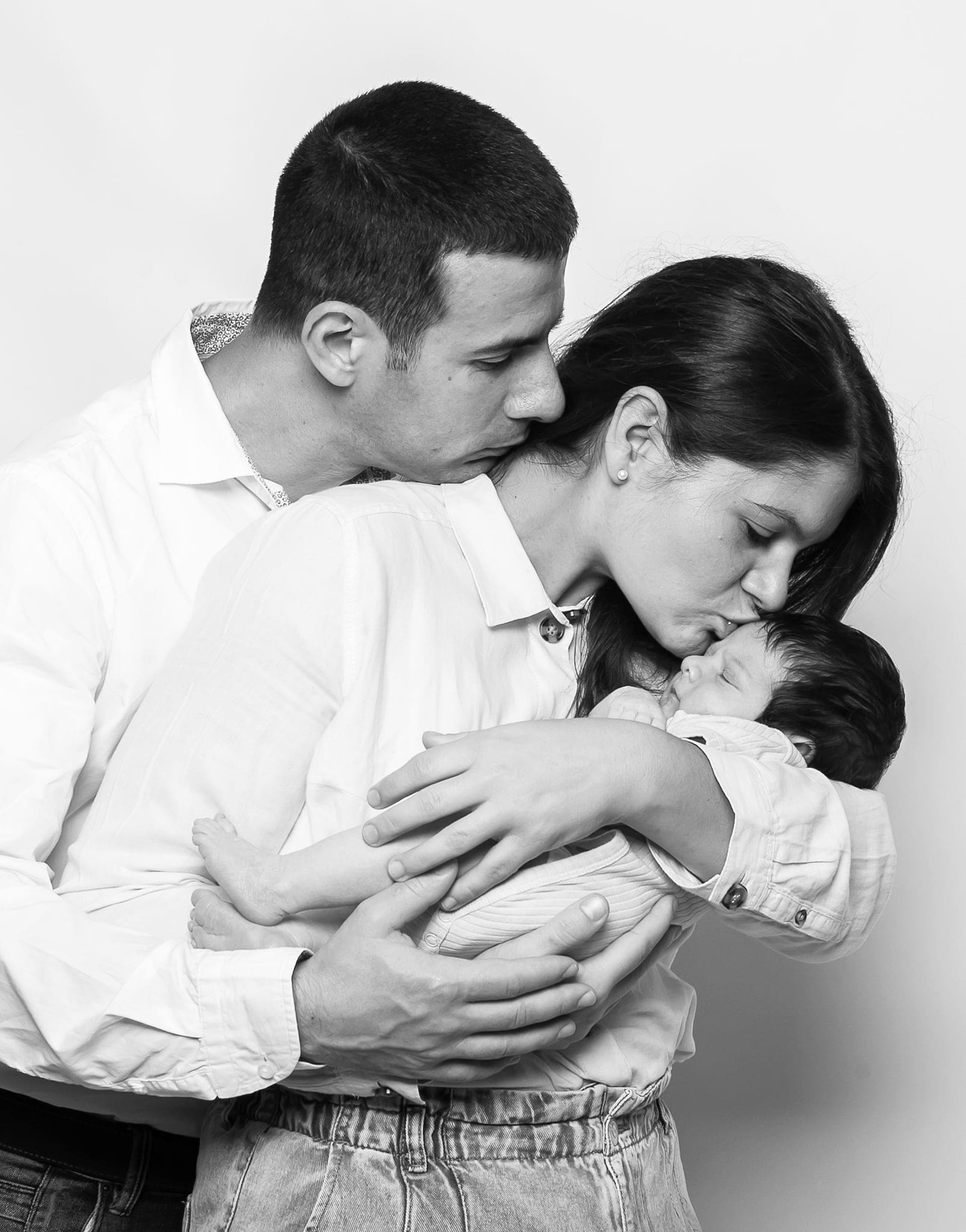 Mamma e papà abbracciati che baciano il loro neonato tenuto tra le braccia, in bianco e nero su fondo chiaro.