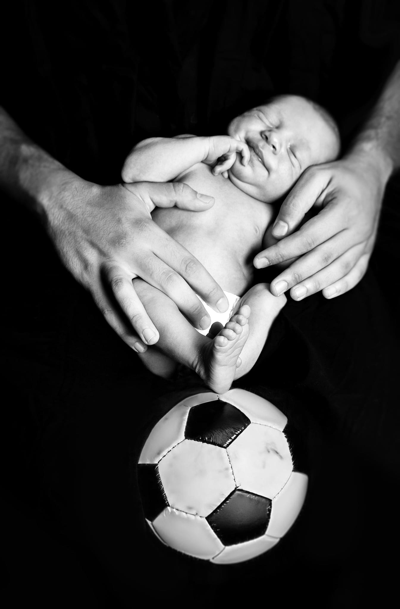 Neonato tenuto da due mani con i piedini che toccano un pallone da calcio, in bianco e nero su fondo nero.