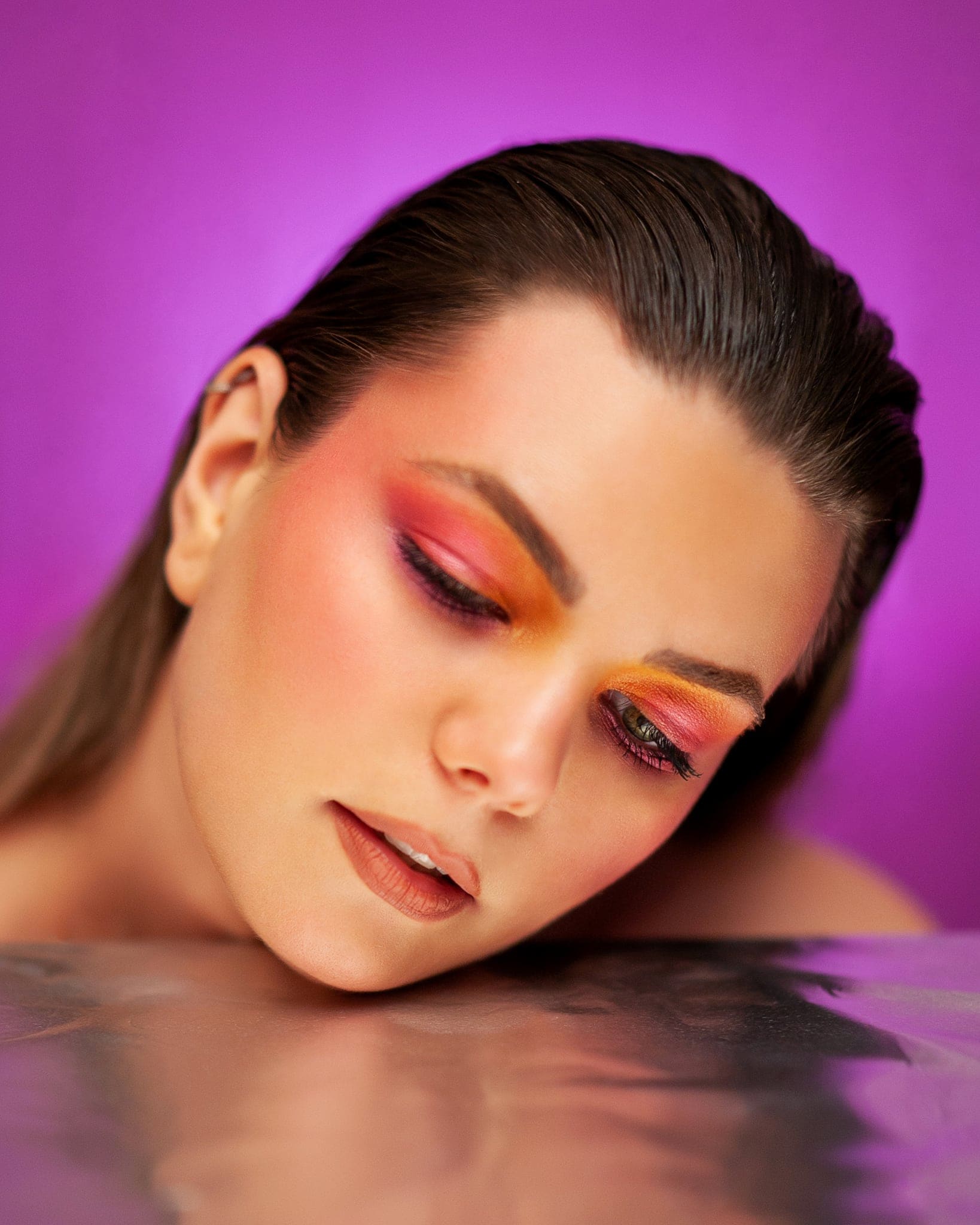 Ragazza con trucco arancio e rosa, capelli tirati indietro e viso appoggiato a una superficie riflettente, su fondo viola