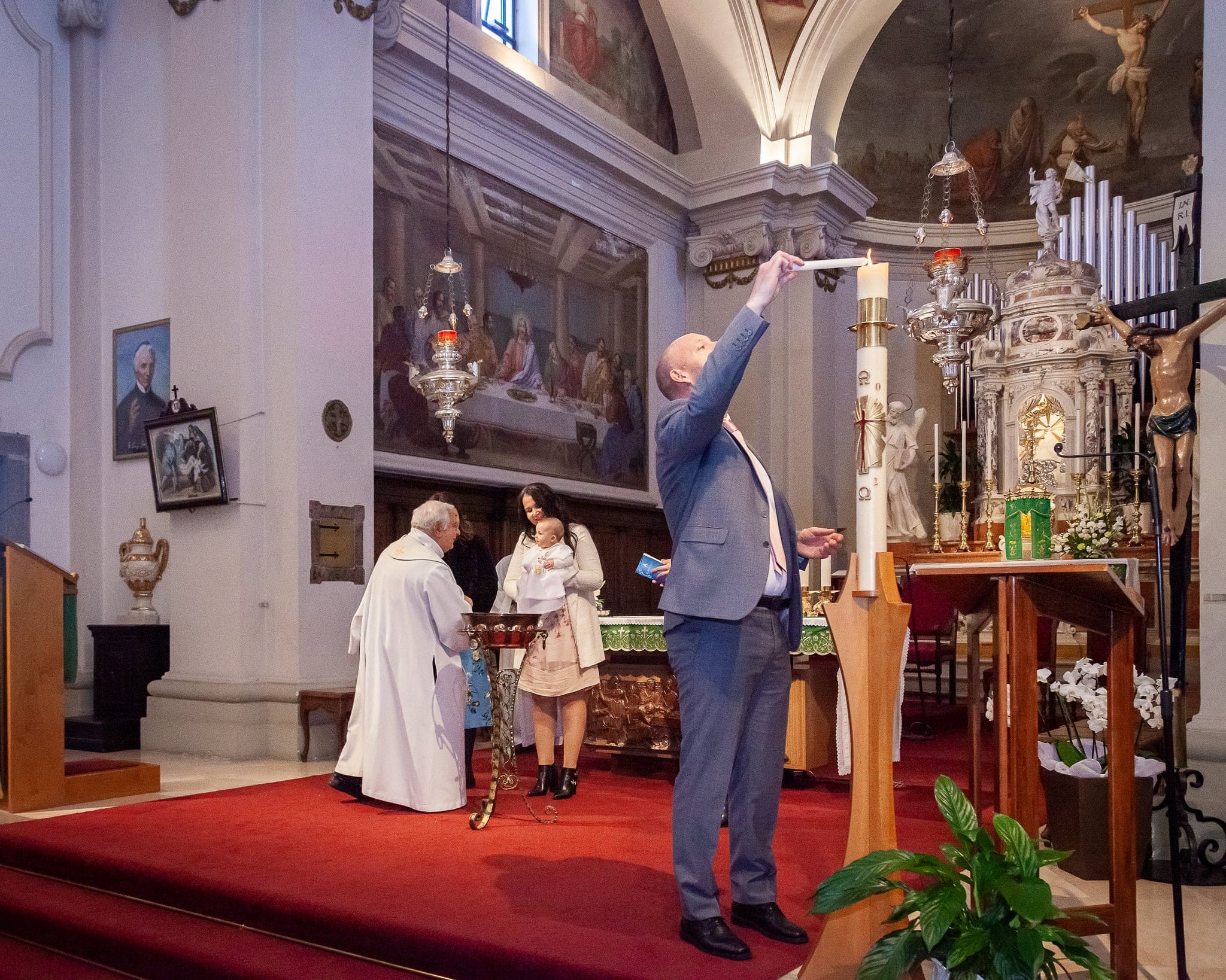 Un uomo accende la candela al cero pasquale vicino all'altare, mentre il sacerdote e la mamma con la bimba attendono sullo sfondo